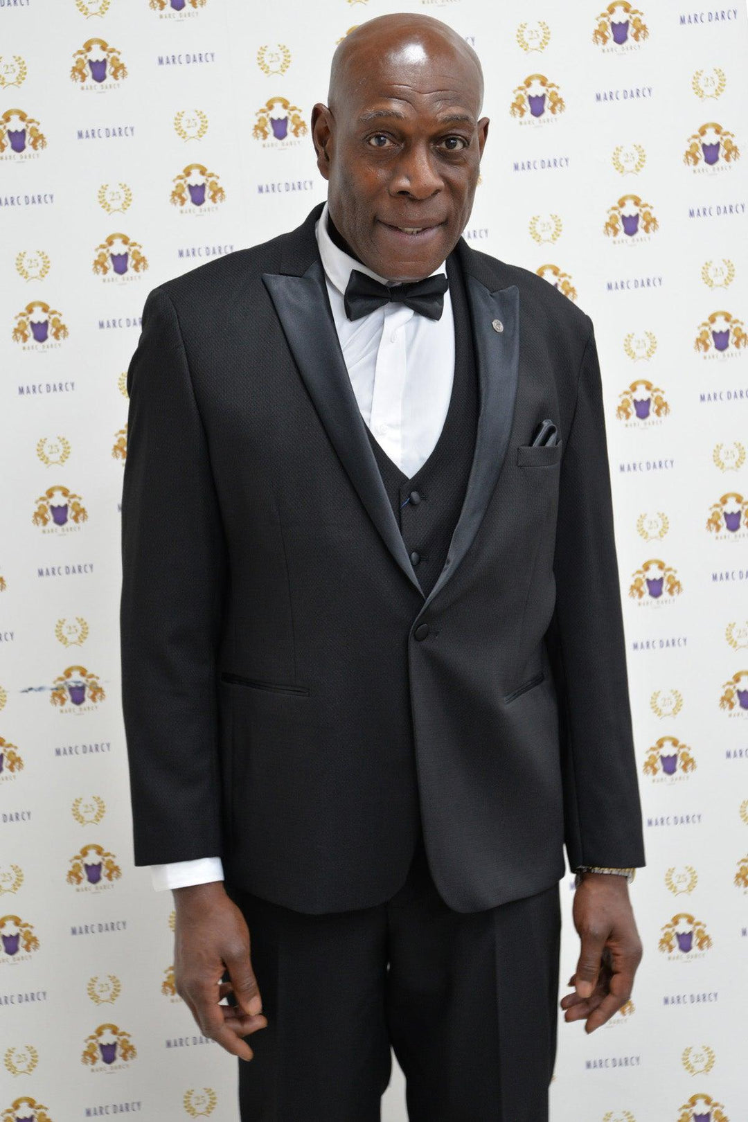 Boxing legend Frank Bruno wears the marcdarcy DALTON Black Tux Lapel Suit, standing before a gold and purple logo backdrop, looking at the camera with a slight smile.