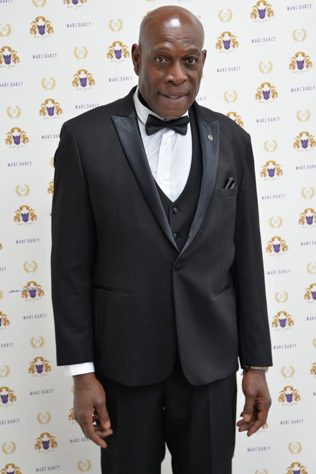 Frank Bruno wears the DALTON Black Tux Lapel Suit by marcdarcy, posing in front of a branded step-and-repeat with gold laurel designs—a sophisticated choice for black-tie events.