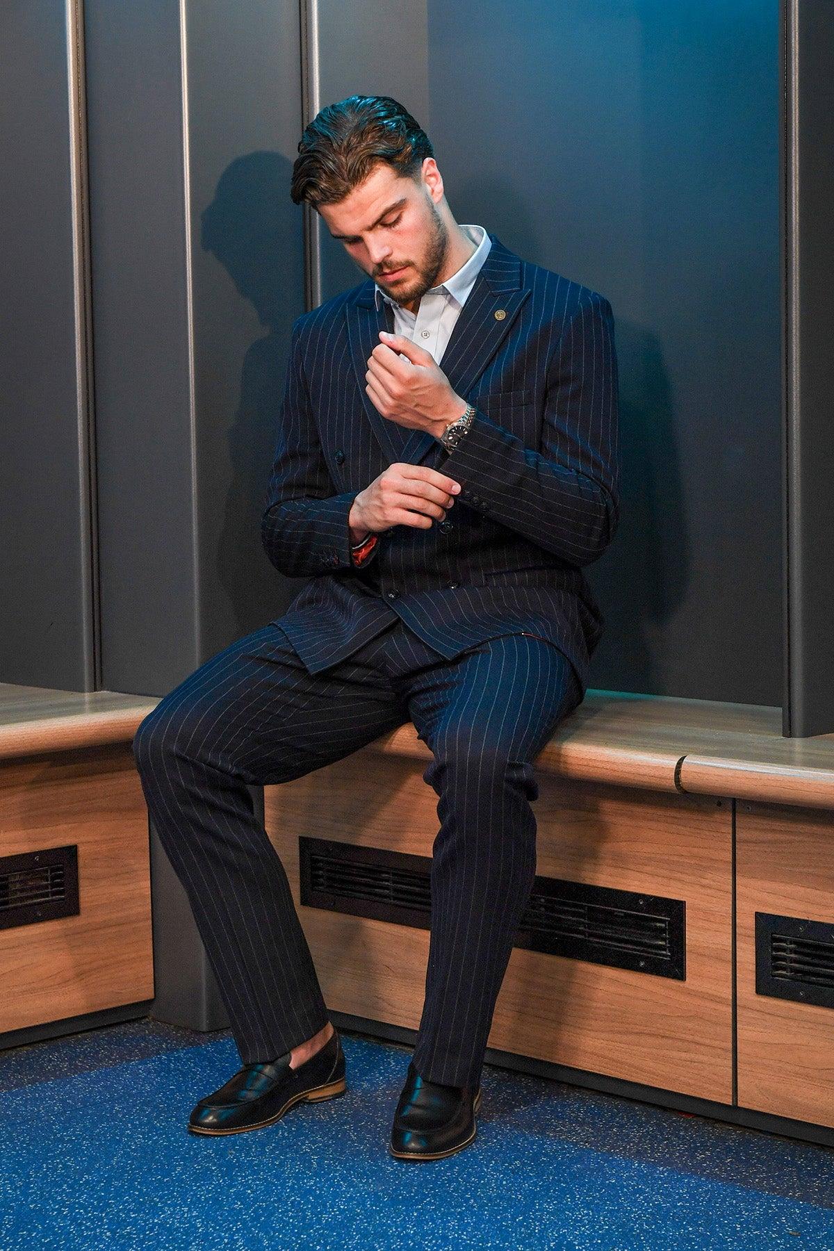A man in the Marc Darcy Cardiff City F.C. Collection ROCCO Navy Pinstripe Double Breasted Suit, as worn by Ollie Tanner, adjusts his cuffs while sitting on a wooden bench.