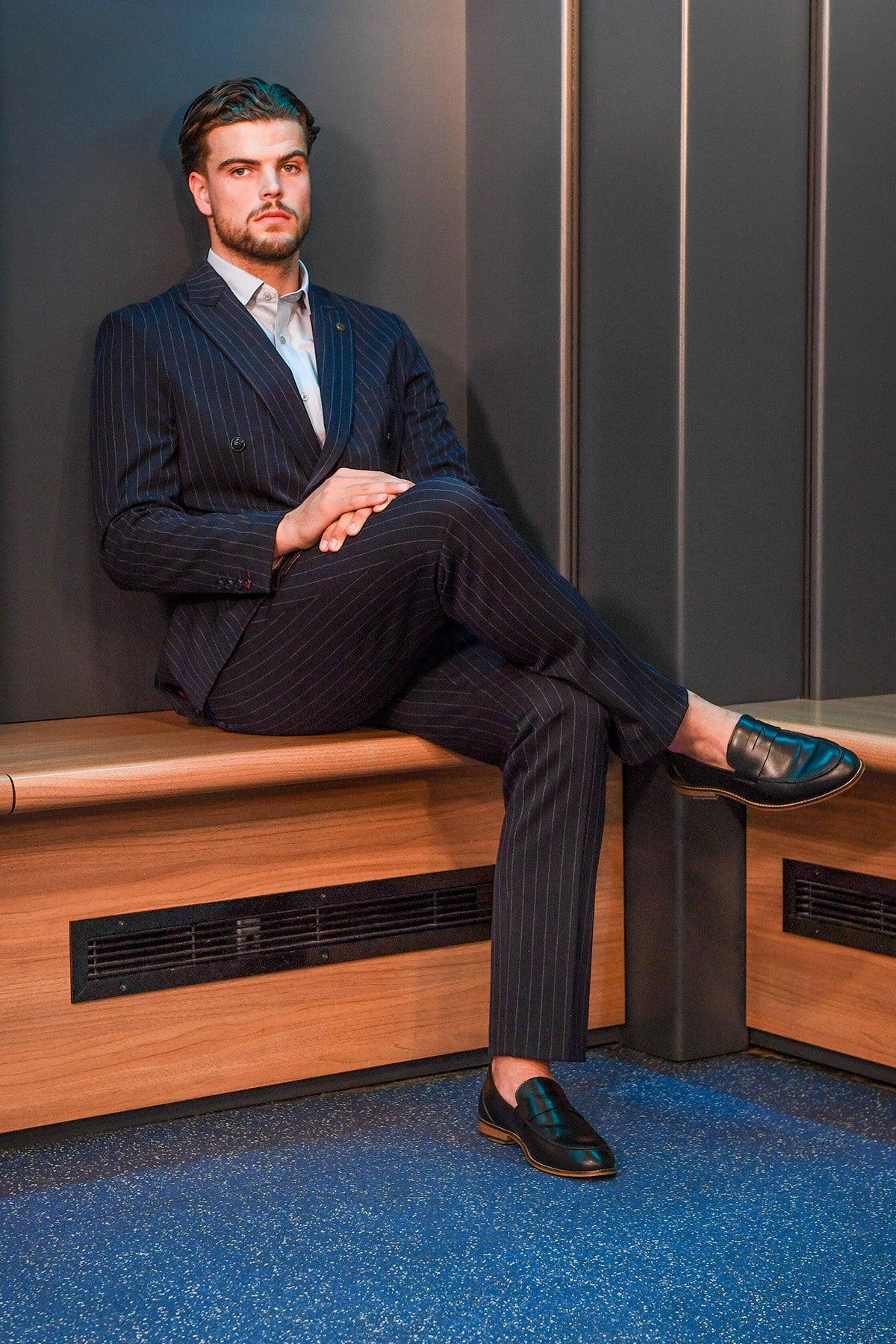 A man wearing the Marc Darcy Cardiff City F.C. Collection – ROCCO Navy Pinstripe Double Breasted Suit as worn by Ollie Tanner sits cross-legged on a bench in a modern room with dark walls and blue speckled flooring.