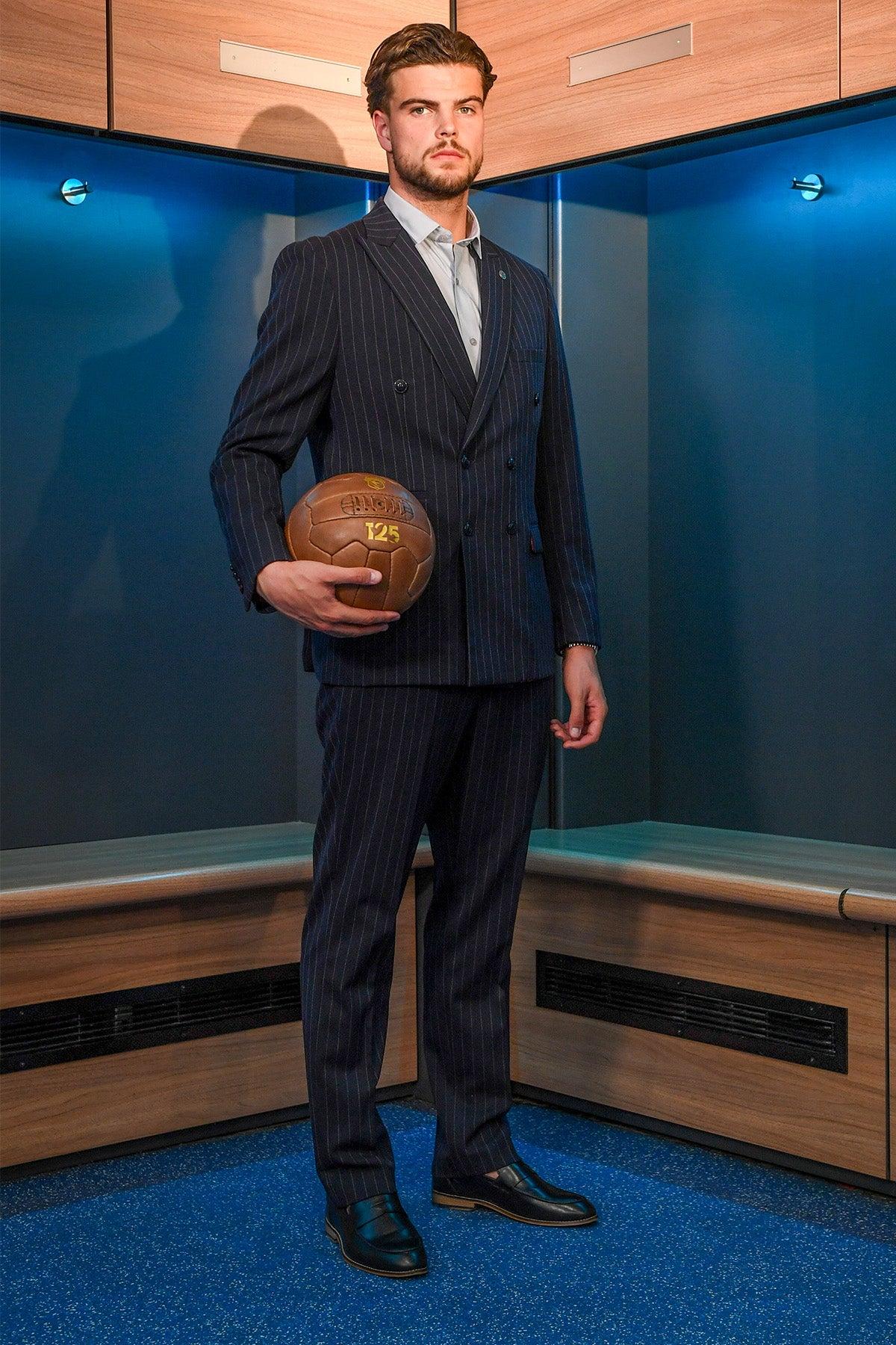 A man with neatly styled hair and a beard stands in a locker room holding a vintage football, wearing the Marc Darcy Cardiff City F.C. Collection - ROCCO Navy Pinstripe Double Breasted Suit, as worn by Ollie Tanner, looking ahead with a serious expression.