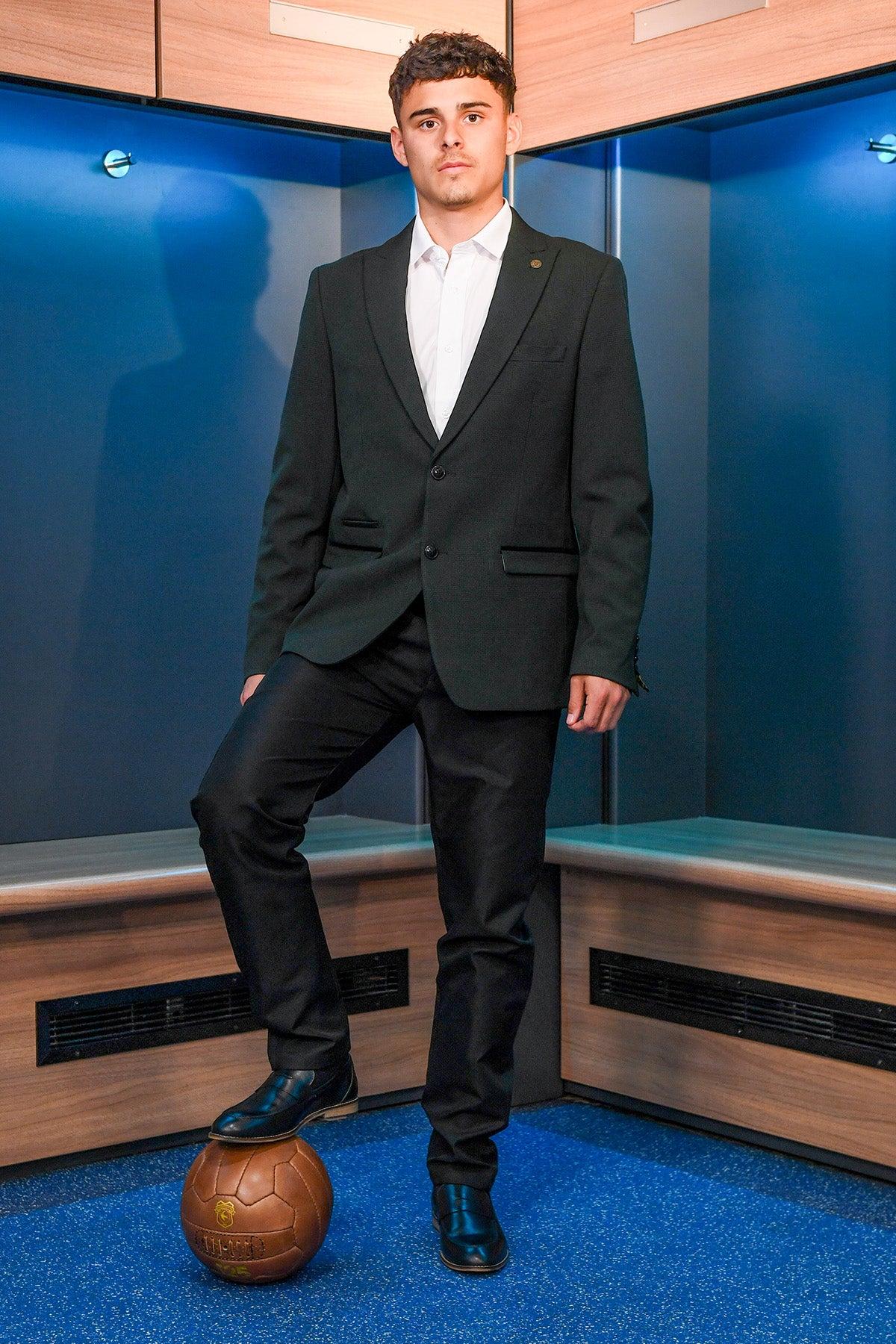 A young man wears the Marc Darcy BROMLEY Olive Green Check Blazer from The Cardiff City F.C. Collection, as worn by Alex Robertson, in a locker room with one foot on a vintage brown soccer ball, looking confidently at the camera.
