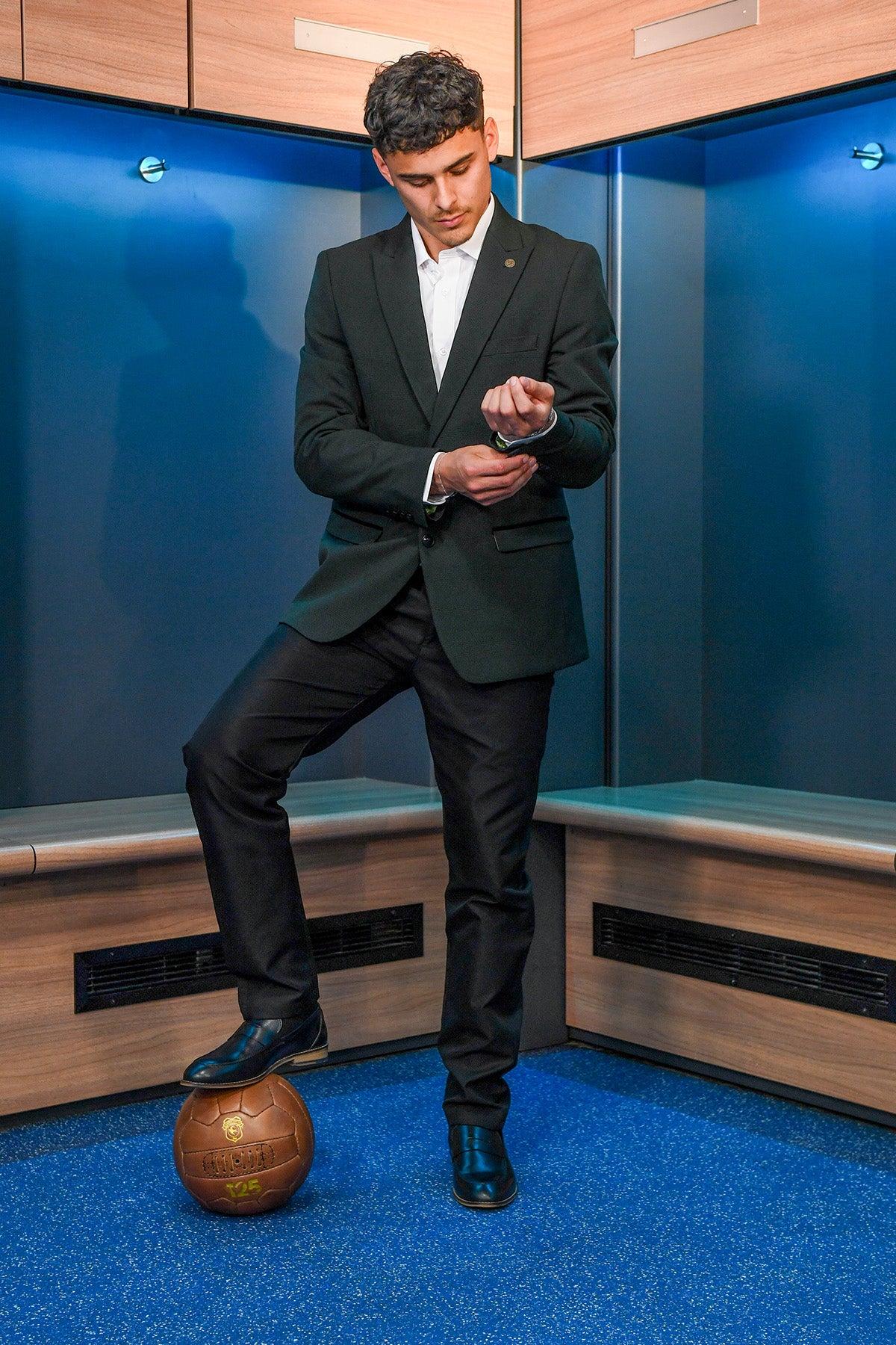 A man wearing the Marc Darcy BROMLEY Olive Green Check Blazer from The Cardiff City F.C. Collection, as worn by Alex Robertson, stands in a locker room with blue floors and wooden benches, adjusting his cufflink with one foot on a vintage brown soccer ball.