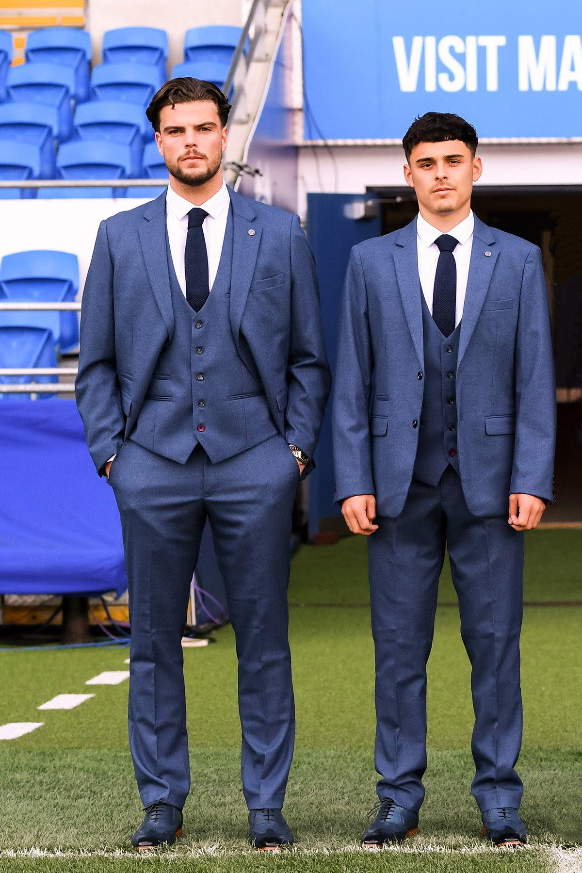 Two men stand side by side on a sports field, both wearing the Marc Darcy Official Cardiff City F.C. Club Suit—ASHLEY Navy Blue Three Piece Suit—with slim fit tailoring, posing formally before blue stadium seating.