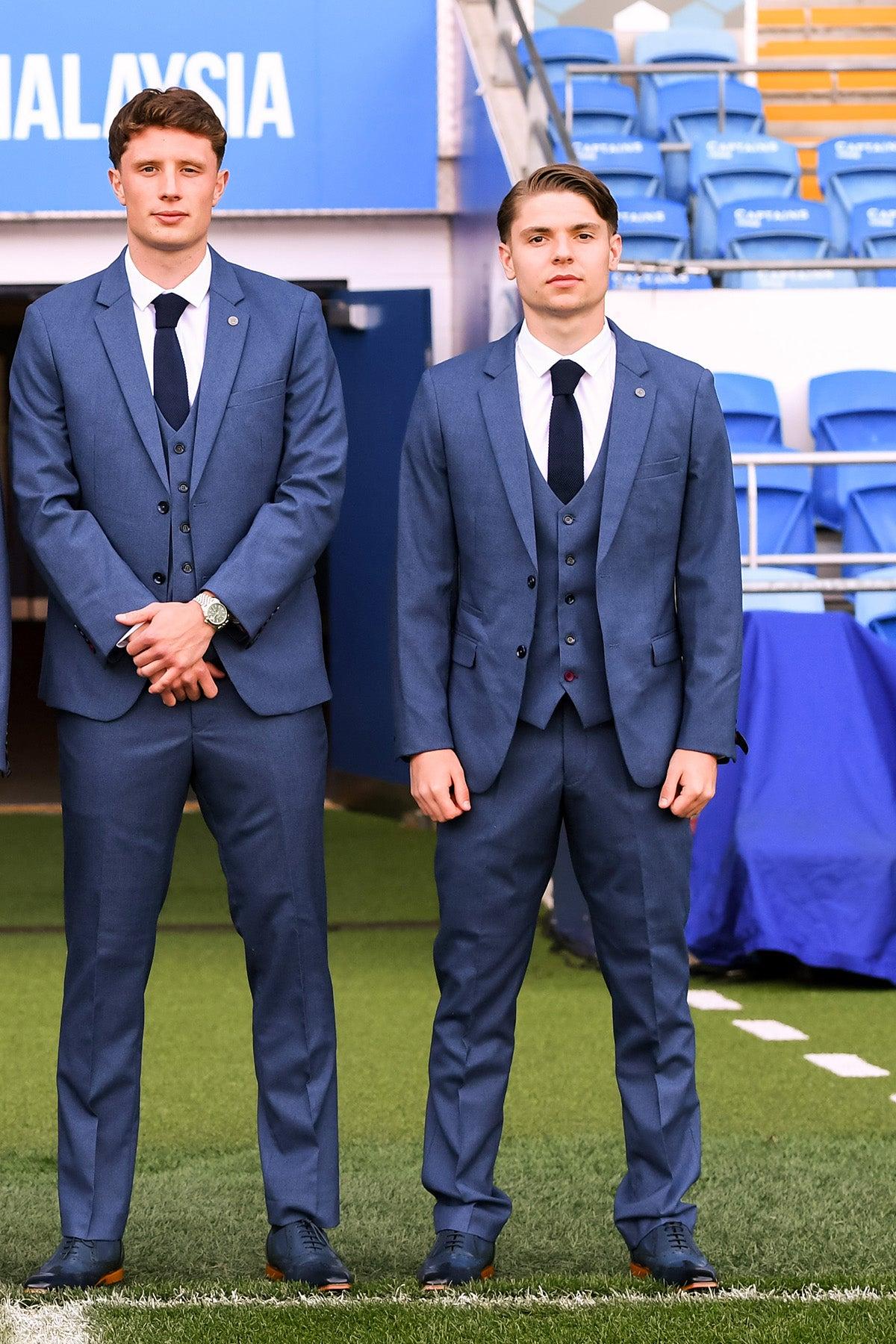 Two young men in matching Marc Darcy Official Cardiff City F.C. Club Suits (ASHLEY Navy Blue Three Piece) stand on a grassy field in front of blue stadium seats.
