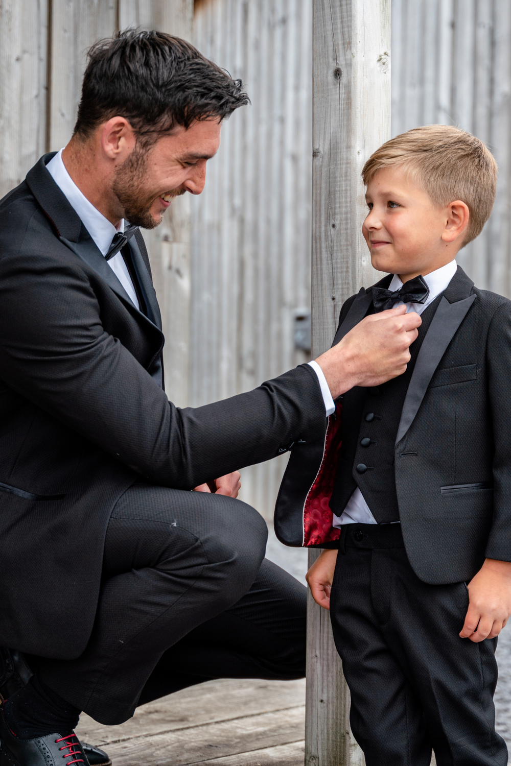 A man and boy, both smiling in marcdarcy's Matching Father & Son | DALTON - Black Tux Three Piece Suit, kneel by a wooden structure against a neutral background as the man adjusts the boy's bow tie.