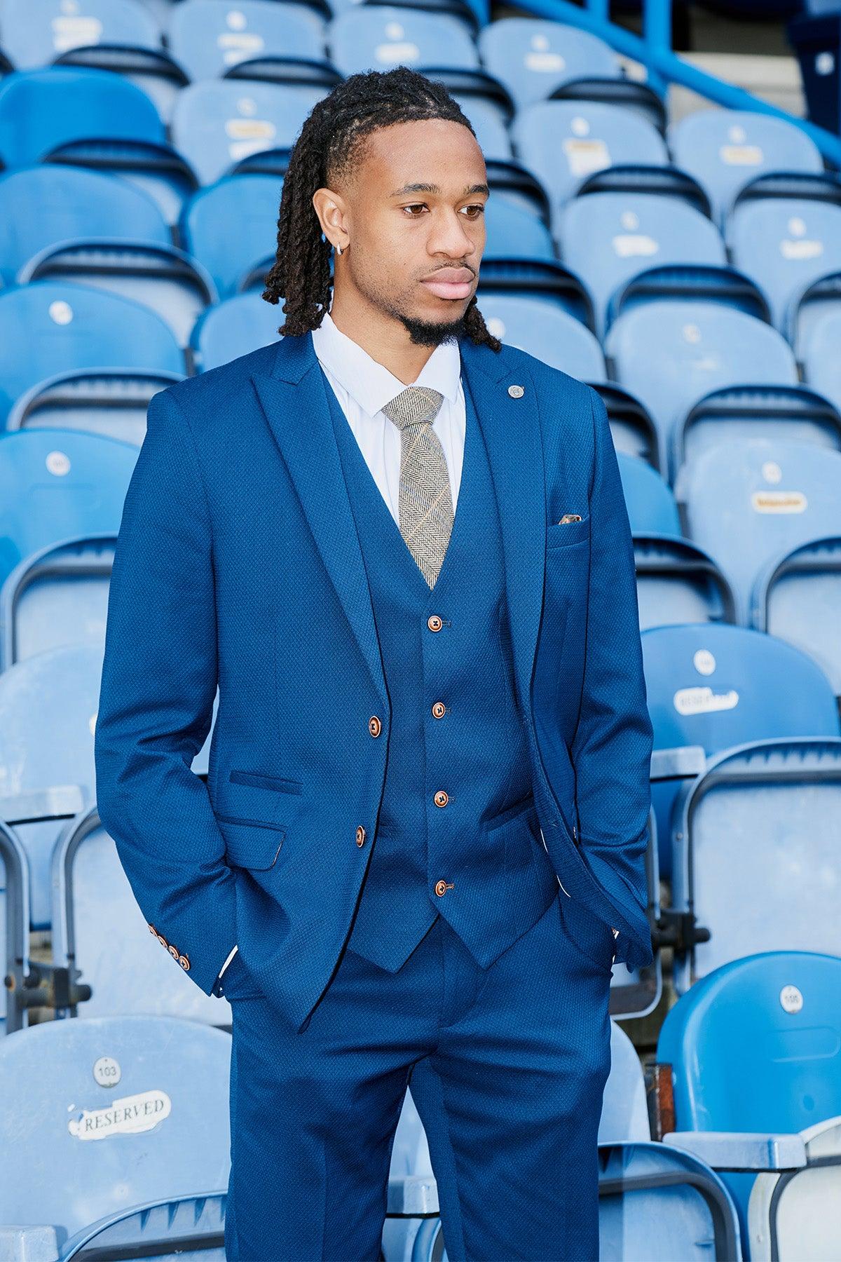 A man in a marcdarcy Huddersfield Town AFC | MAX Royal Blue Suit As Worn By David Kasumu stands with his hands in his pockets in front of blue stadium seats.