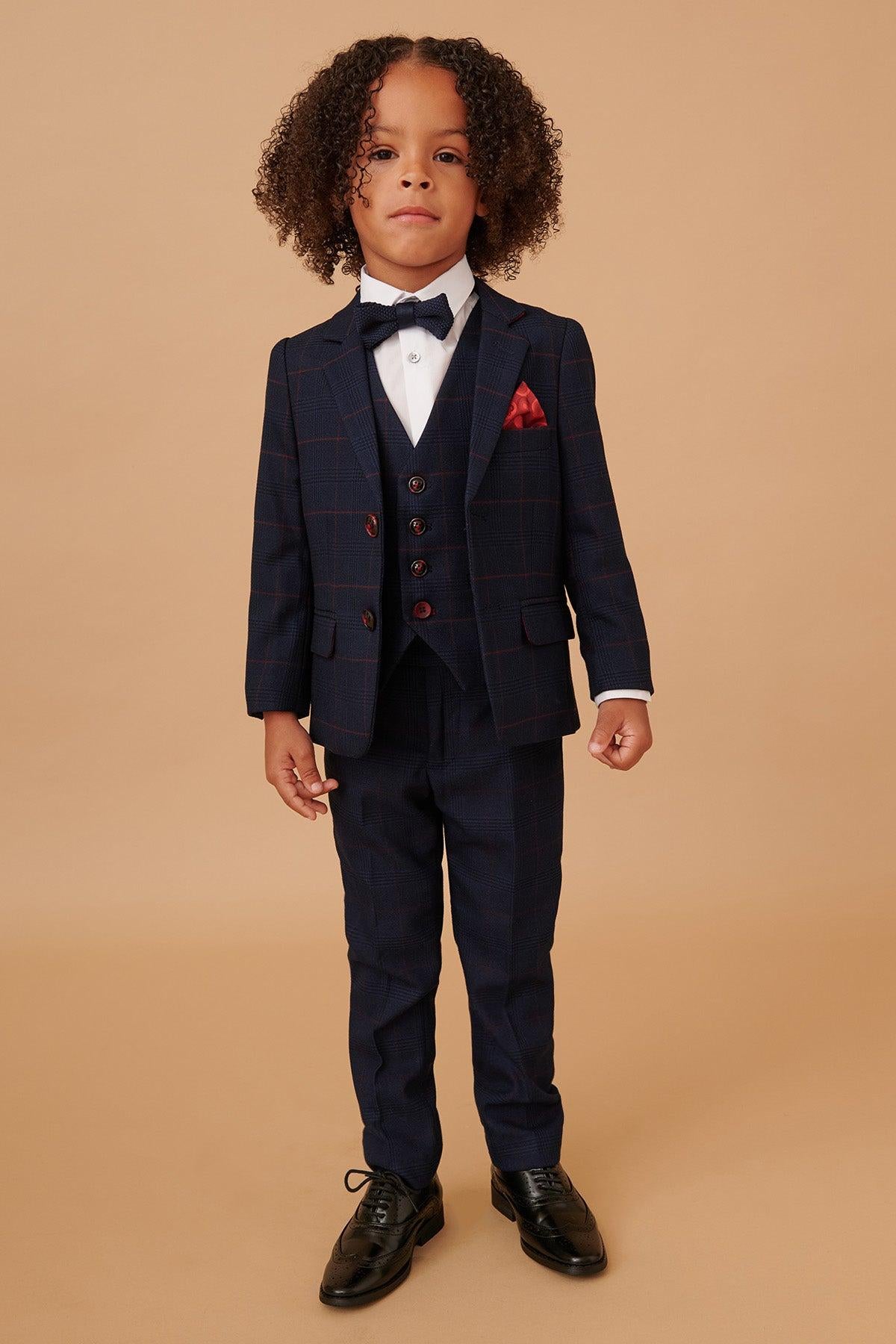 A young boy with curly hair wears the Marc Darcy EDINSON Children's Navy Wine Check Print Three Piece Suit, featuring a white shirt, black bowtie, red pocket square, and black dress shoes.