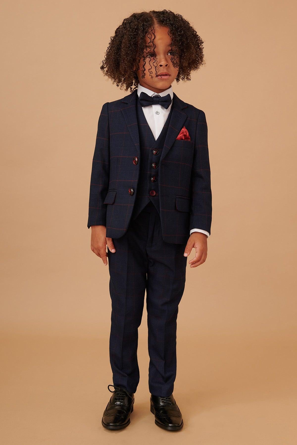 A young child with curly hair wears the EDINSON - Children's Navy Wine Check Print Three Piece Suit by Marc Darcy, paired with a white shirt, bow tie, red pocket square, and black dress shoes against a plain beige background.