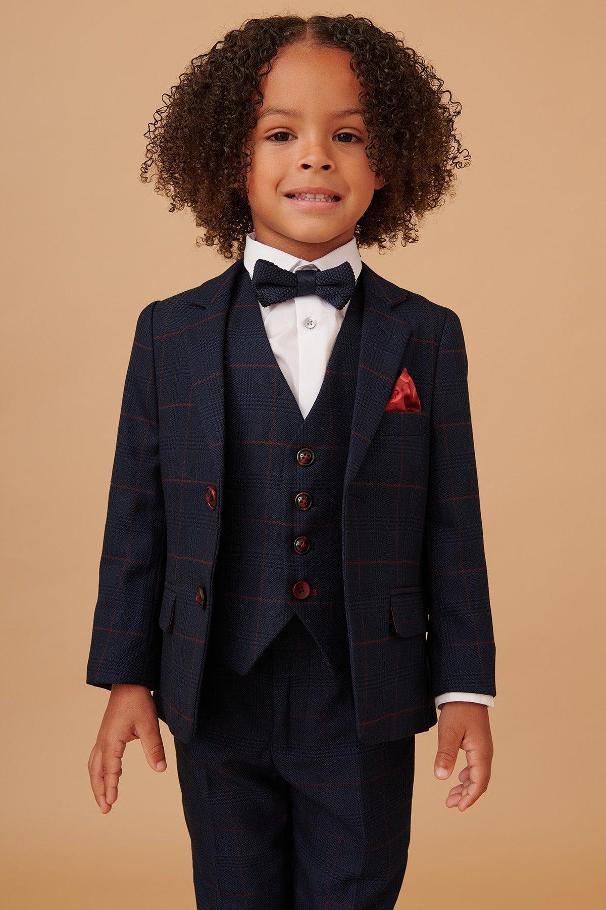 A young child smiles in the Marc Darcy EDINSON Childrens Navy Wine Check Print Three Piece Suit—featuring a matching vest, white shirt, black bow tie, and red pocket square—perfect for special occasions.