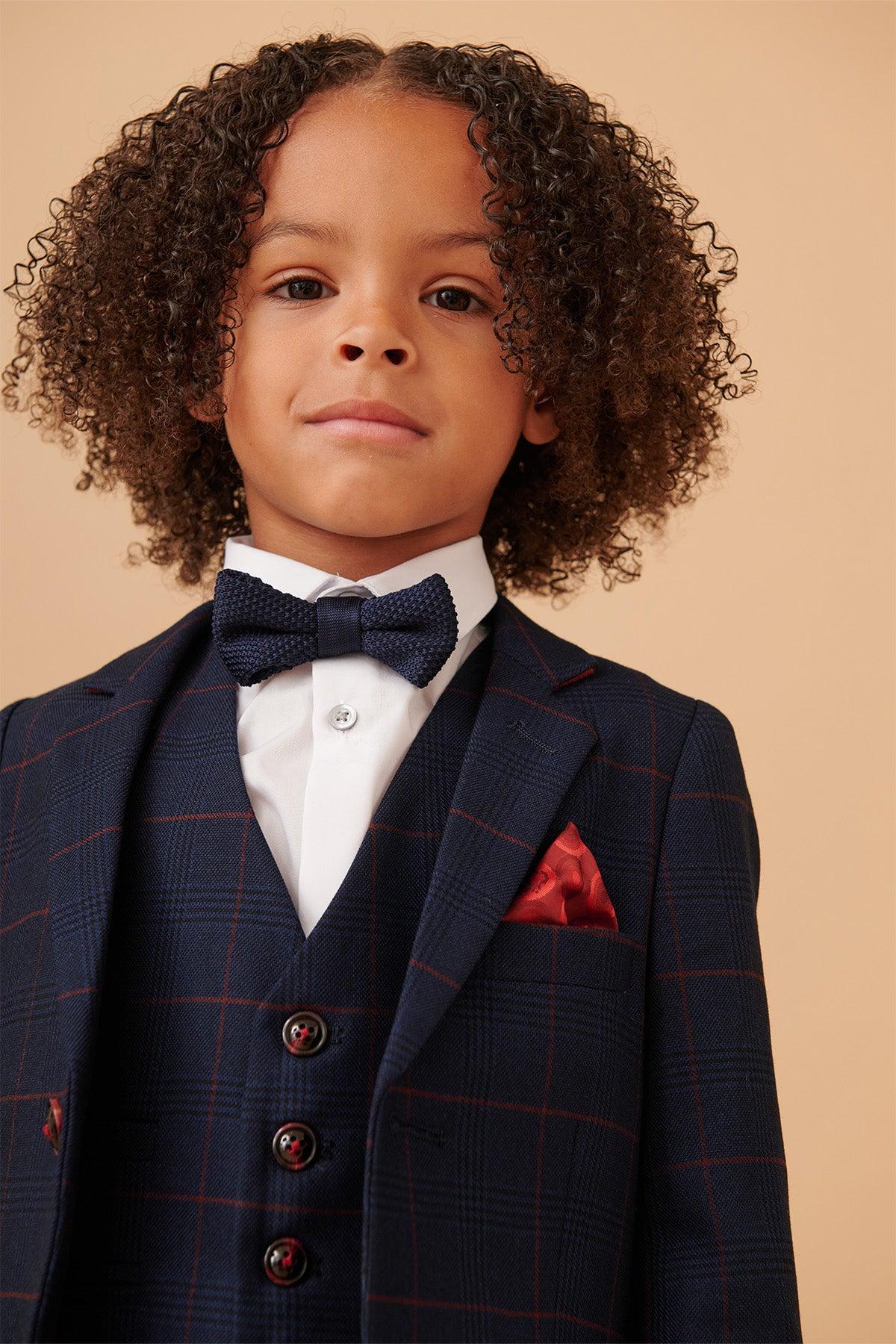 A young child with curly hair wears the Marc Darcy EDINSON Children's Navy Wine Check Print Three Piece Suit, featuring a white shirt, navy bow tie, and red pocket square, standing against a beige background.