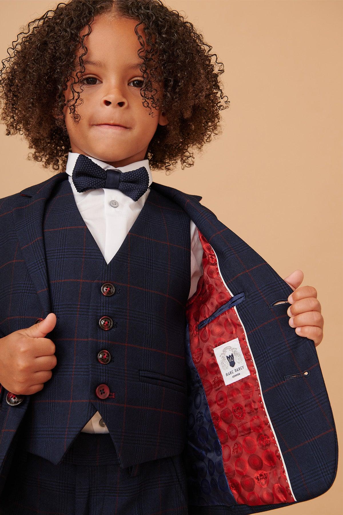 A young child wears the Marc Darcy EDINSON children's navy wine check print three piece suit, paired with a white shirt and bow tie, holding open the jacket to reveal its red patterned lining and inner label. The background is light brown.