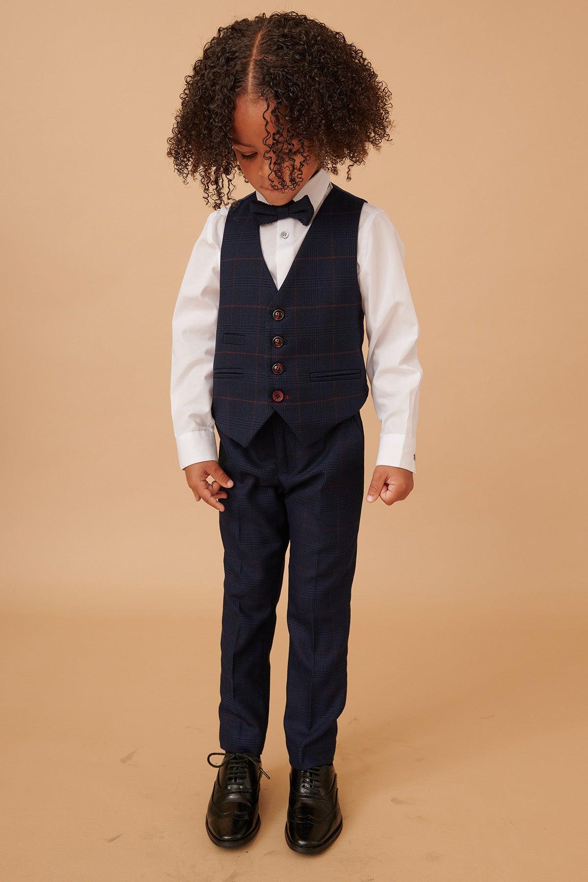 A young child with curly hair wears the Marc Darcy EDINSON Children's Navy Wine Check Print Three Piece Suit, complete with vest, white shirt, bow tie, and black shoes, standing against a plain beige background and looking down.