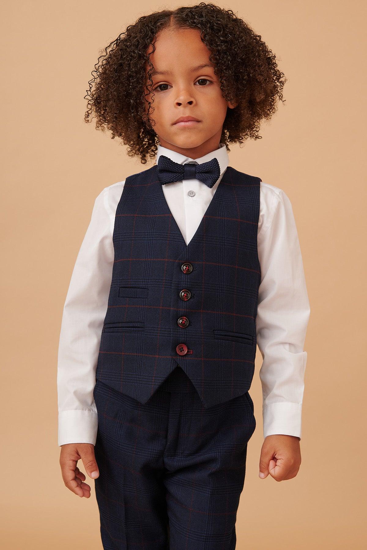 A young boy with curly hair wears the Marc Darcy EDINSON Children's Navy Wine Check Print Three Piece Suit—white shirt, navy check vest and trousers, and a navy bow tie—standing against a beige background.