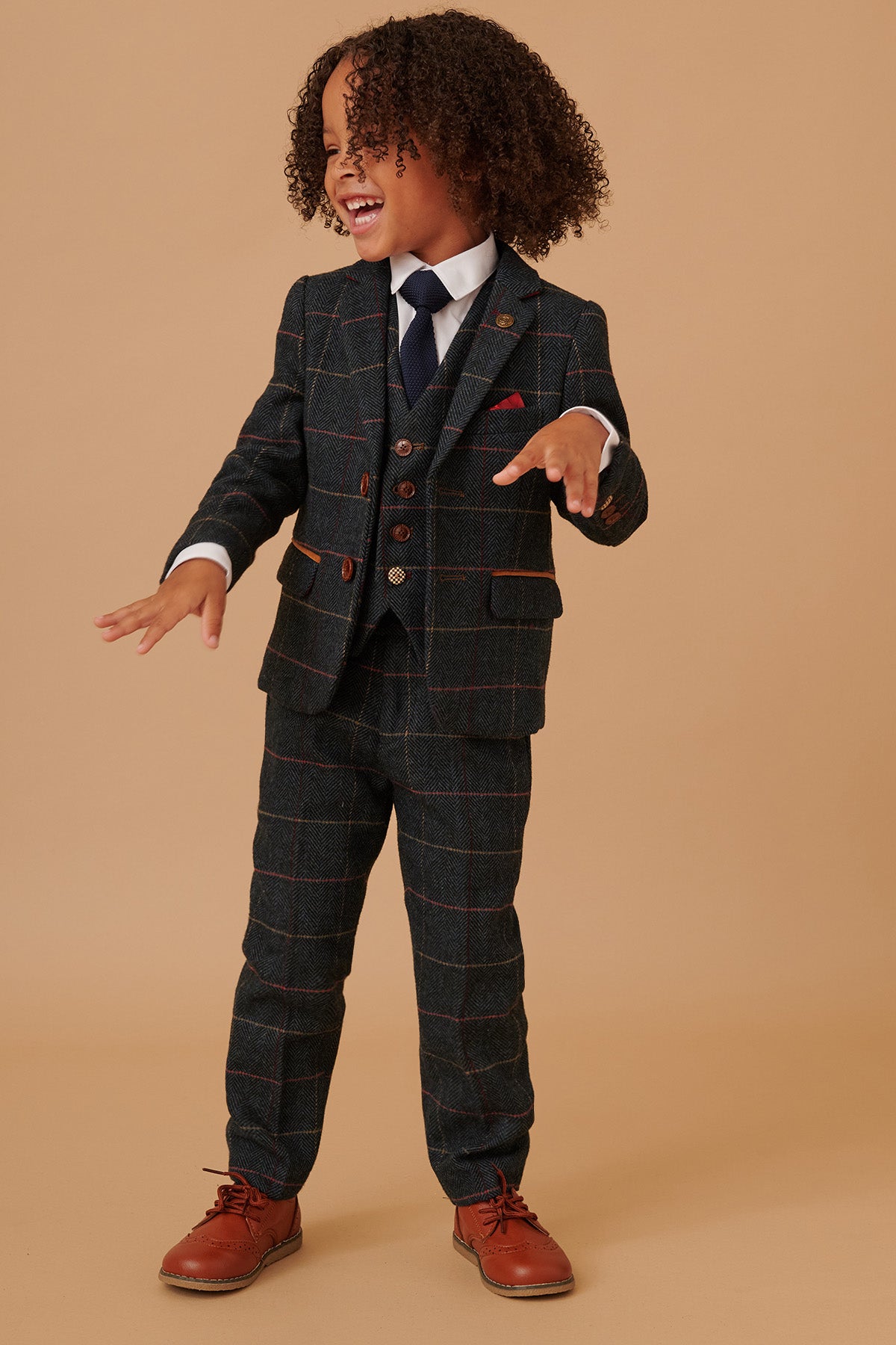 A young boy with curly hair smiles and gestures in a marcdarcy ETON Childrens Navy Blue Tweed Check Three Piece Suit, paired with a white shirt, boys gold paisley print tie, and brown shoes against a beige background.