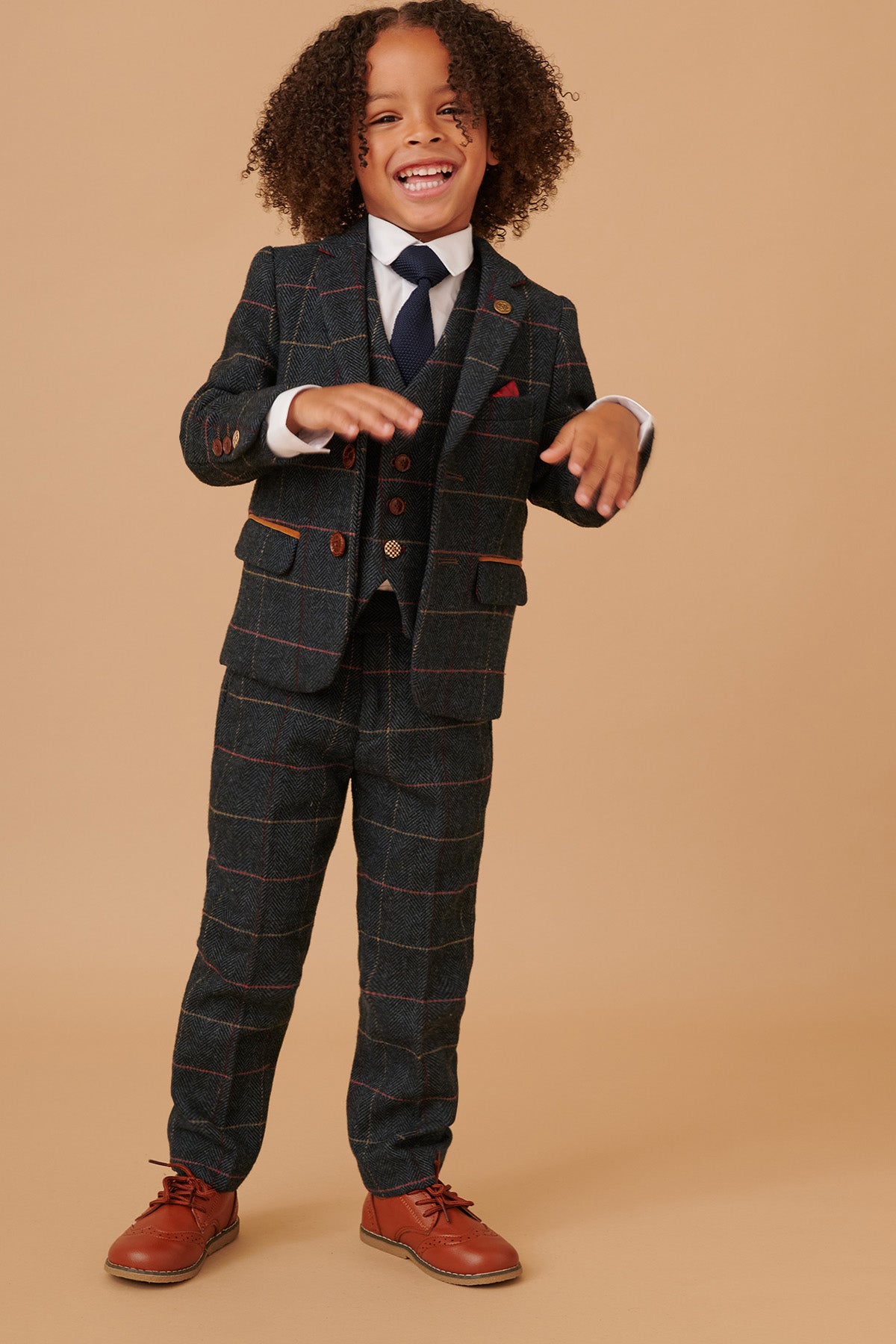 A young child with curly hair smiles while wearing the marcdarcy ETON Childrens Navy Blue Tweed Check Three Piece Suit, paired with a white shirt and brown shoes, standing against a plain beige background.