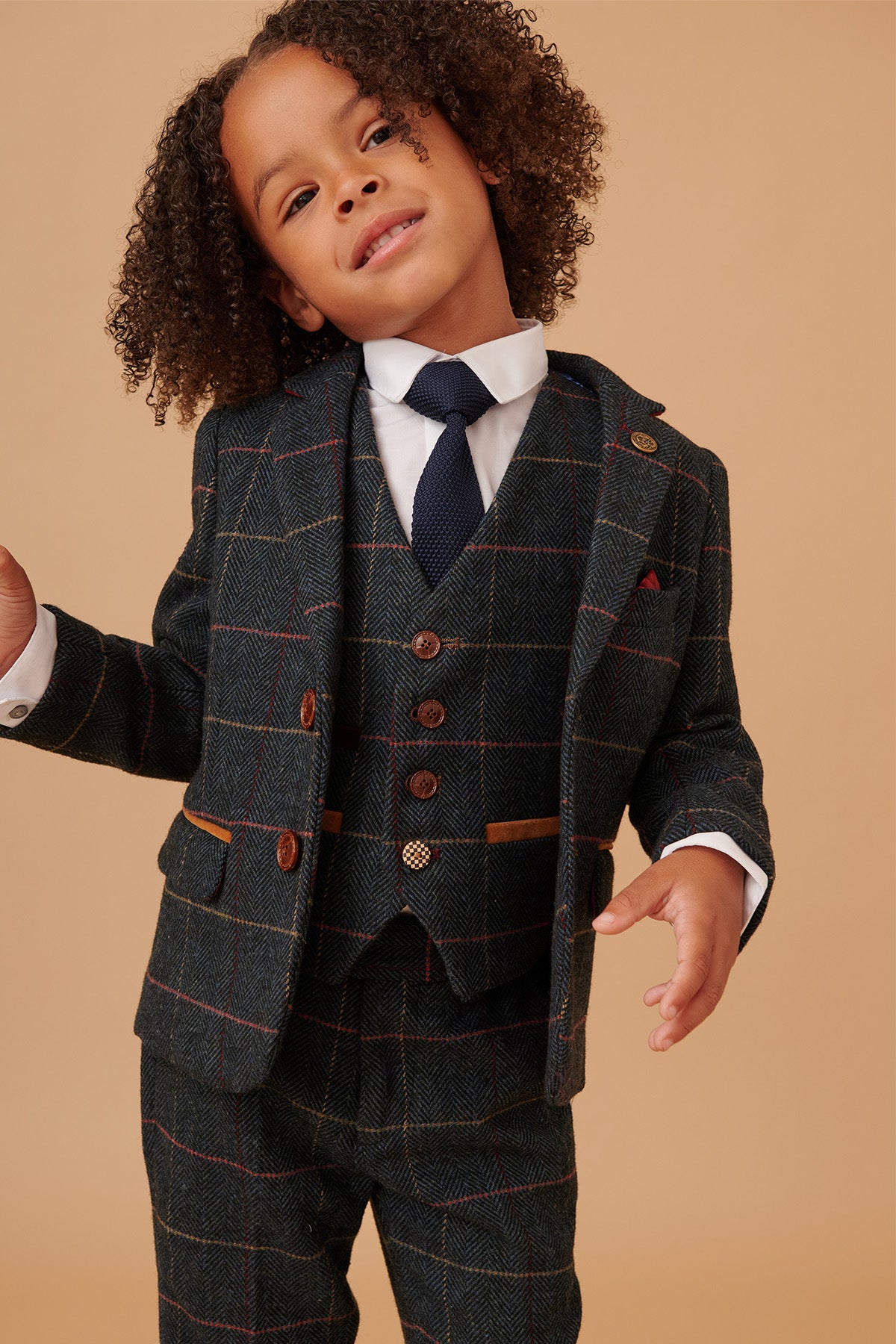 A young child with curly hair smiles, wearing the marcdarcy ETON Childrens Navy Blue Tweed Check Three Piece Suit, featuring a vest and white shirt, with a boys gold paisley print tie against a beige background.