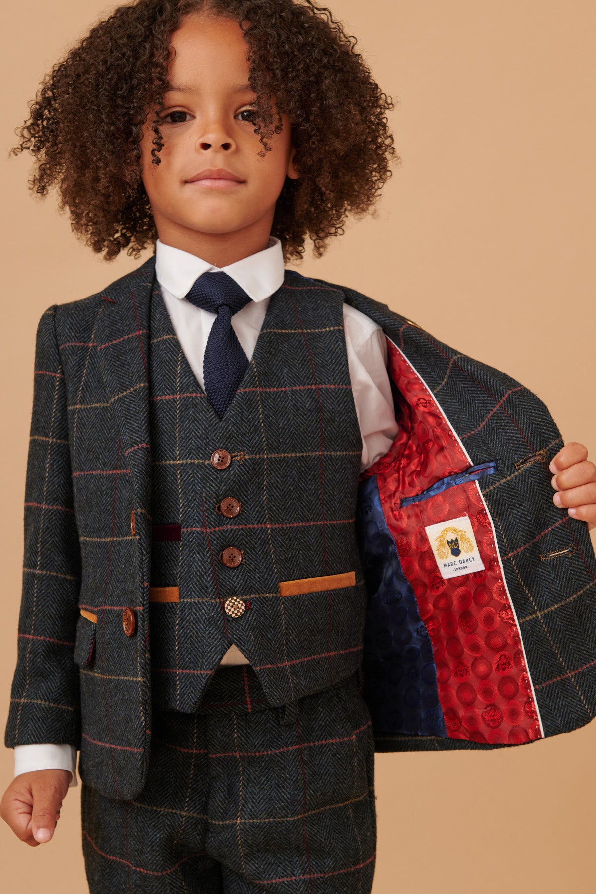 A young boy with curly hair wears the marcdarcy ETON Children's Navy Blue Tweed Check Three Piece Suit, featuring a vest, tie, and a bright red patterned lining inside the jacket.