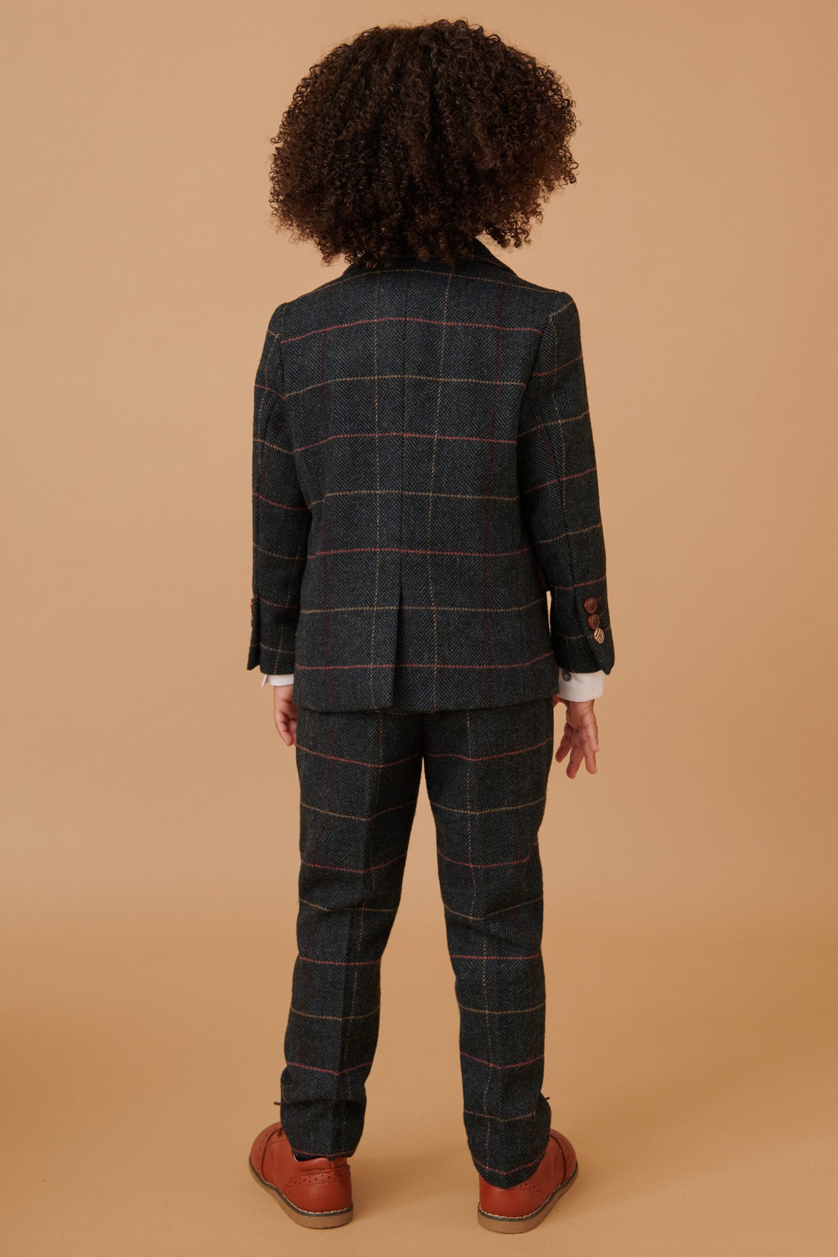 A child faces away from the camera, wearing the marcdarcy ETON Children's Navy Blue Tweed Check Three Piece Suit, which features a jacket and pants, paired with brown shoes.