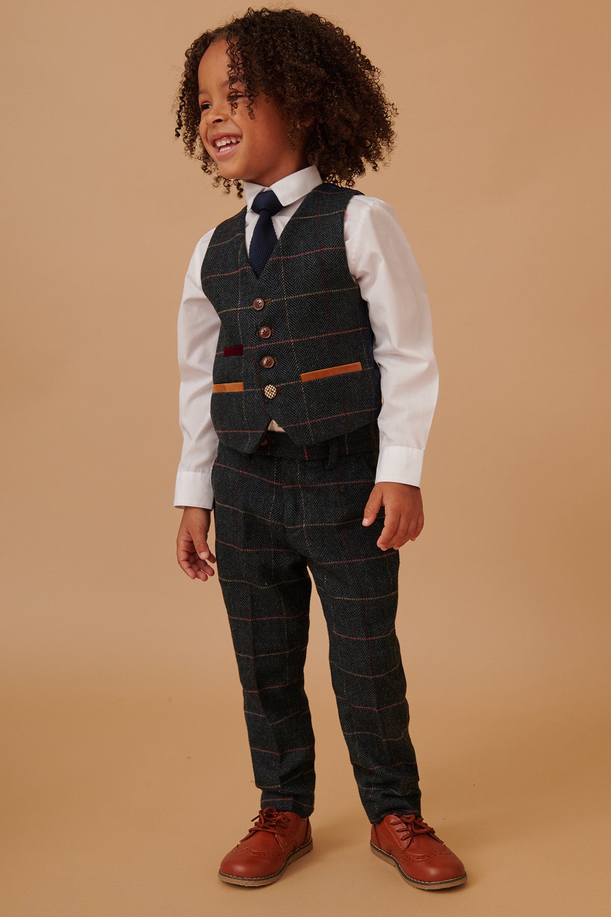 A young boy with curly hair smiles in the marcdarcy ETON Children’s Navy Blue Tweed Check Three Piece Suit, featuring a vest and matching pants over a white shirt and tie—perfect for coordinating adult and child outfits.