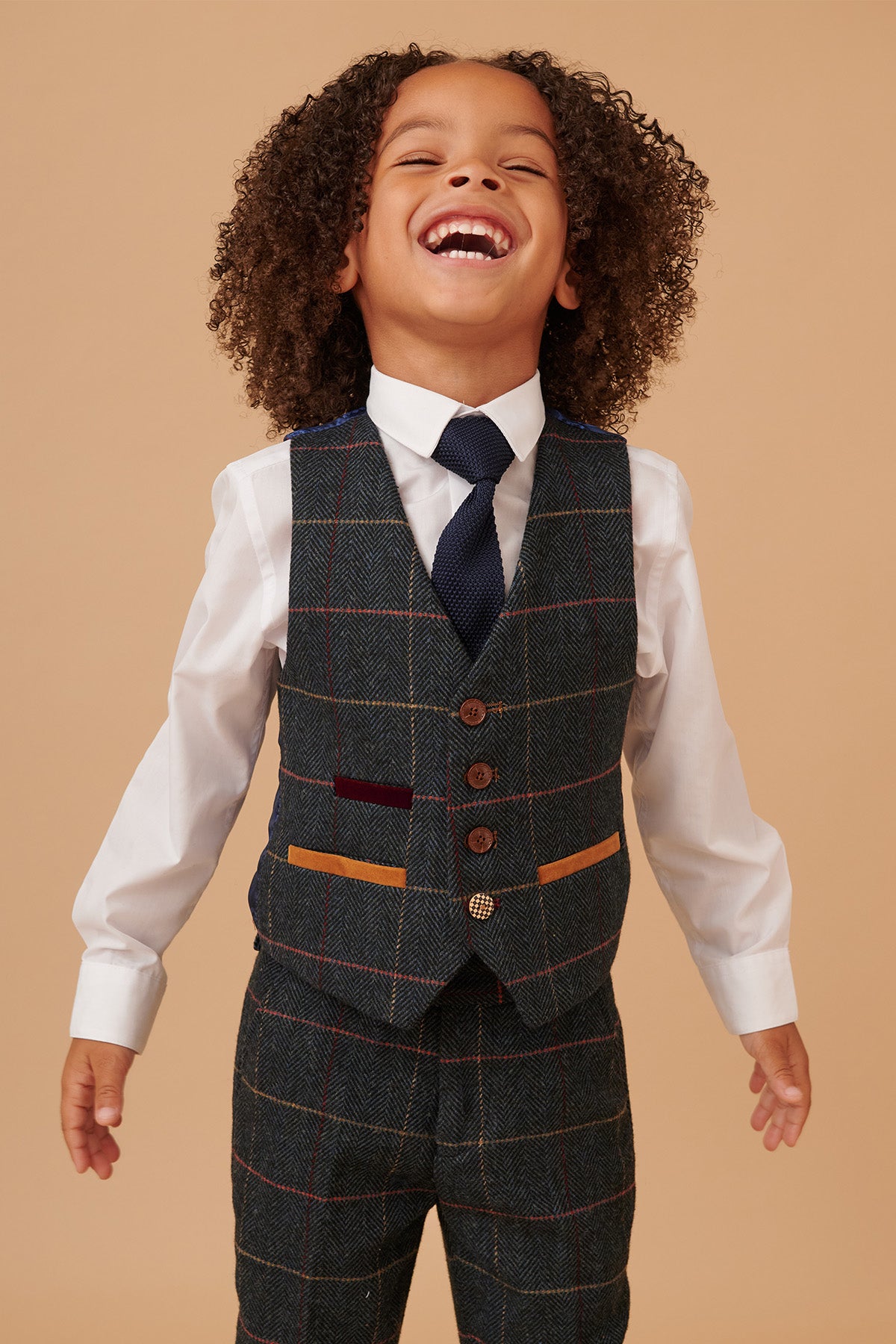 A young boy with curly hair laughs heartily, dressed in the marcdarcy ETON Children's Navy Blue Tweed Check Three Piece Suit.