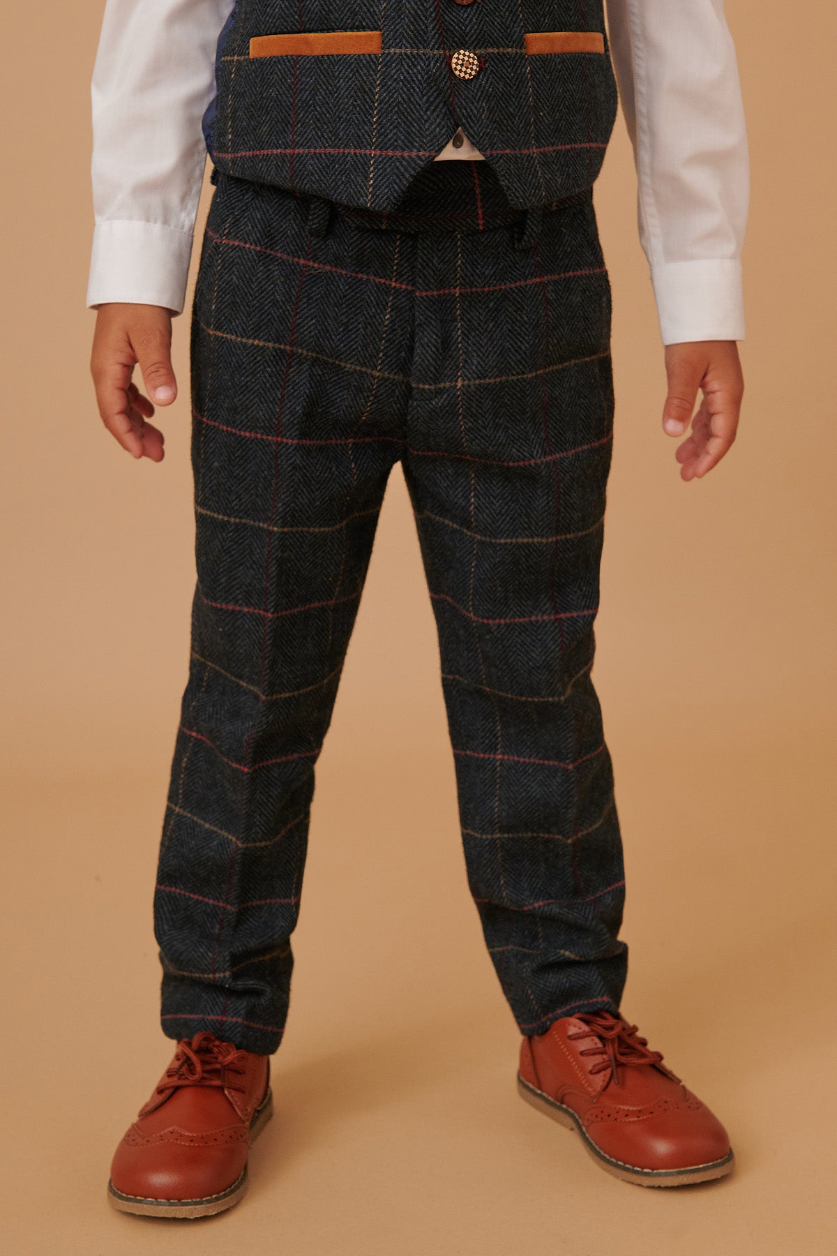 A young child models the marcdarcy ETON - Childrens Navy Blue Tweed Check Three Piece Suit, featuring dress pants and a matching vest, paired with a white shirt and brown dress shoes. The beige background image is cropped from the shoulders down.