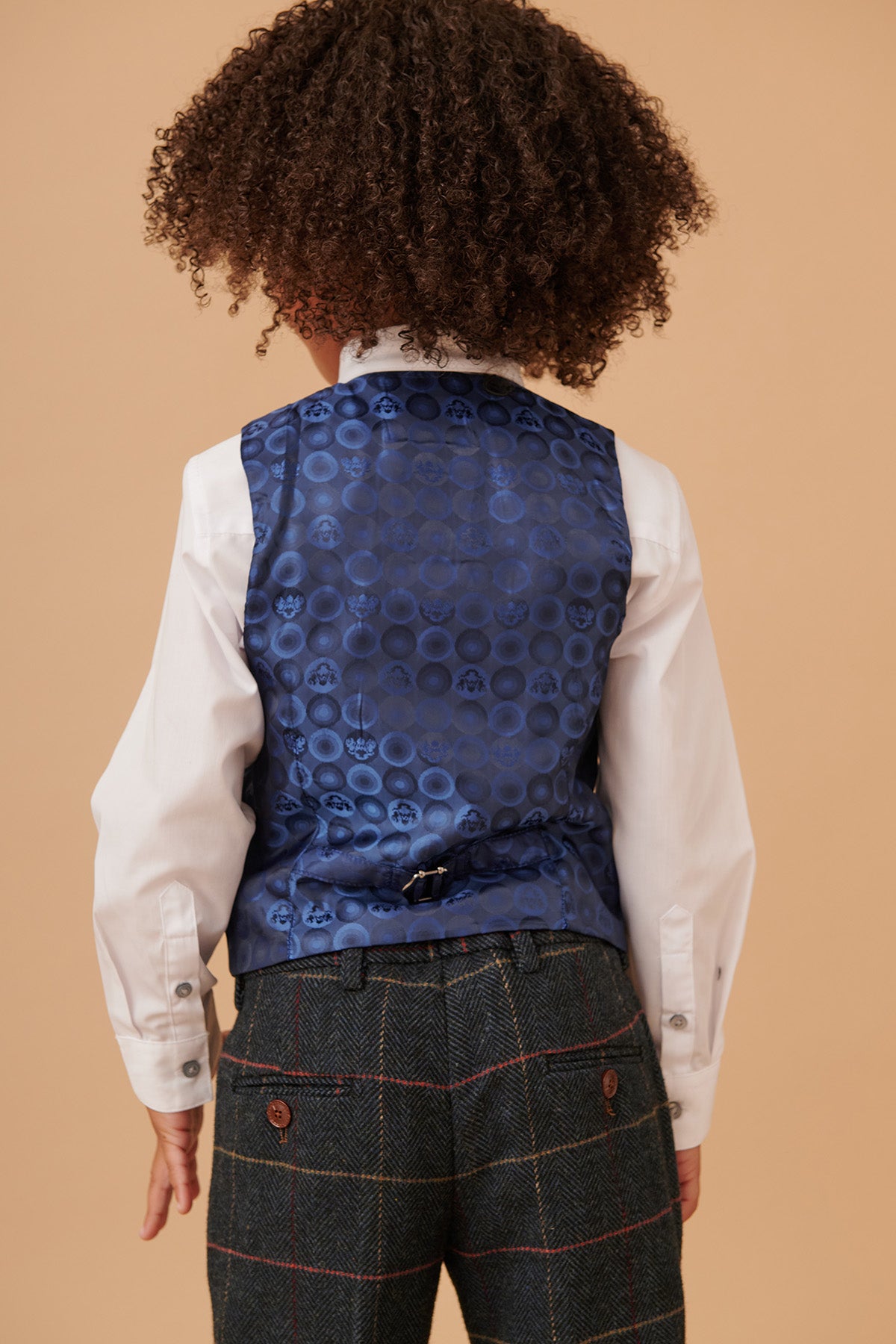 A child with curly hair, viewed from behind, wears the marcdarcy ETON Childrens Navy Blue Tweed Check Three Piece Suit, styled with a white dress shirt and boys gold paisley print tie, standing against a plain beige background.