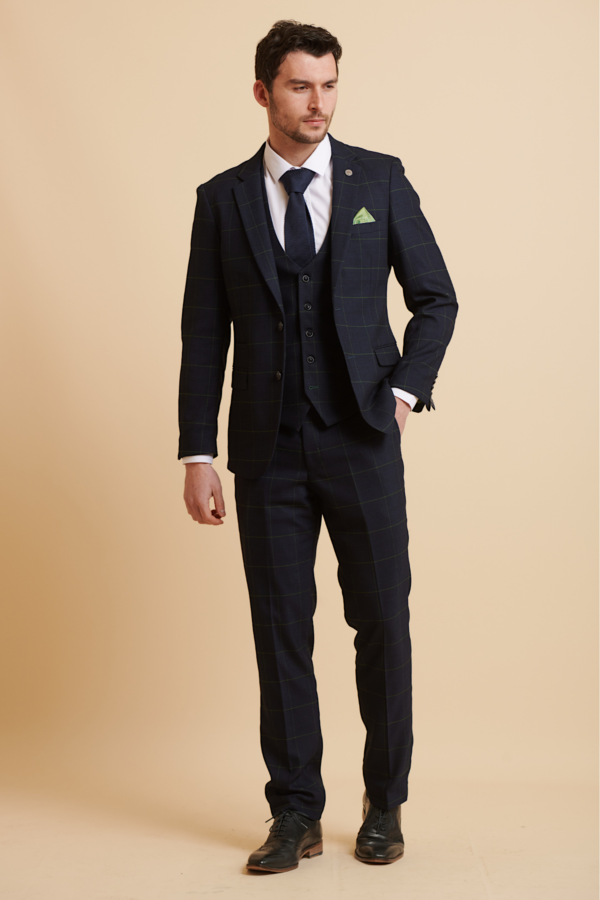 James Tarkowski models The Everton Collection EDINSON Navy Green Check Suit by Marc Darcy against a beige background, styled with a white shirt, navy tie, green pocket square, and black shoes. He has short dark hair, a trimmed beard, and one hand in his pocket.