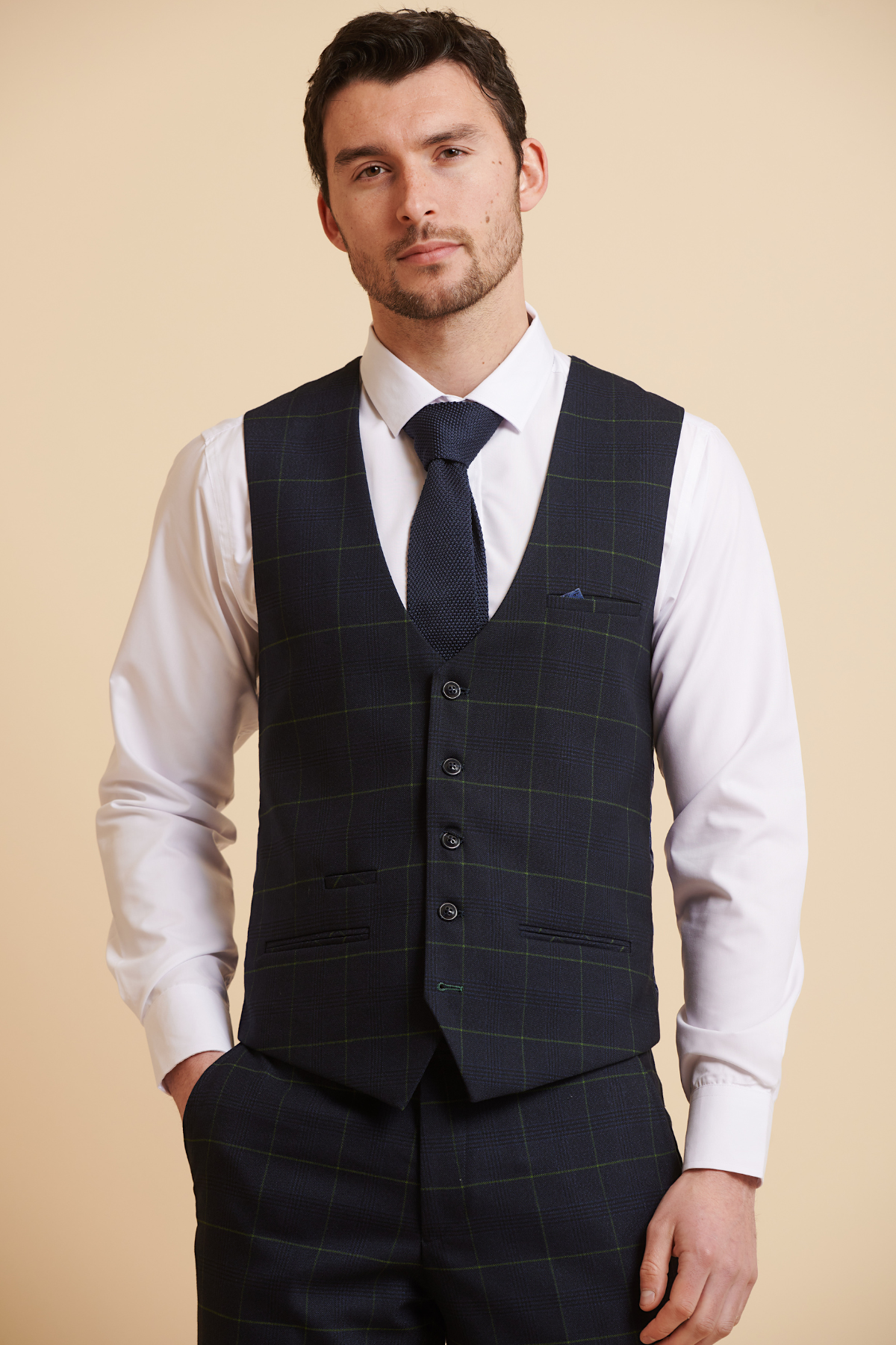 A man in a Marc Darcy EDINSON Navy Green Check Single Breasted Waistcoat, white dress shirt, and dark blue tie stands with one hand in his pocket.