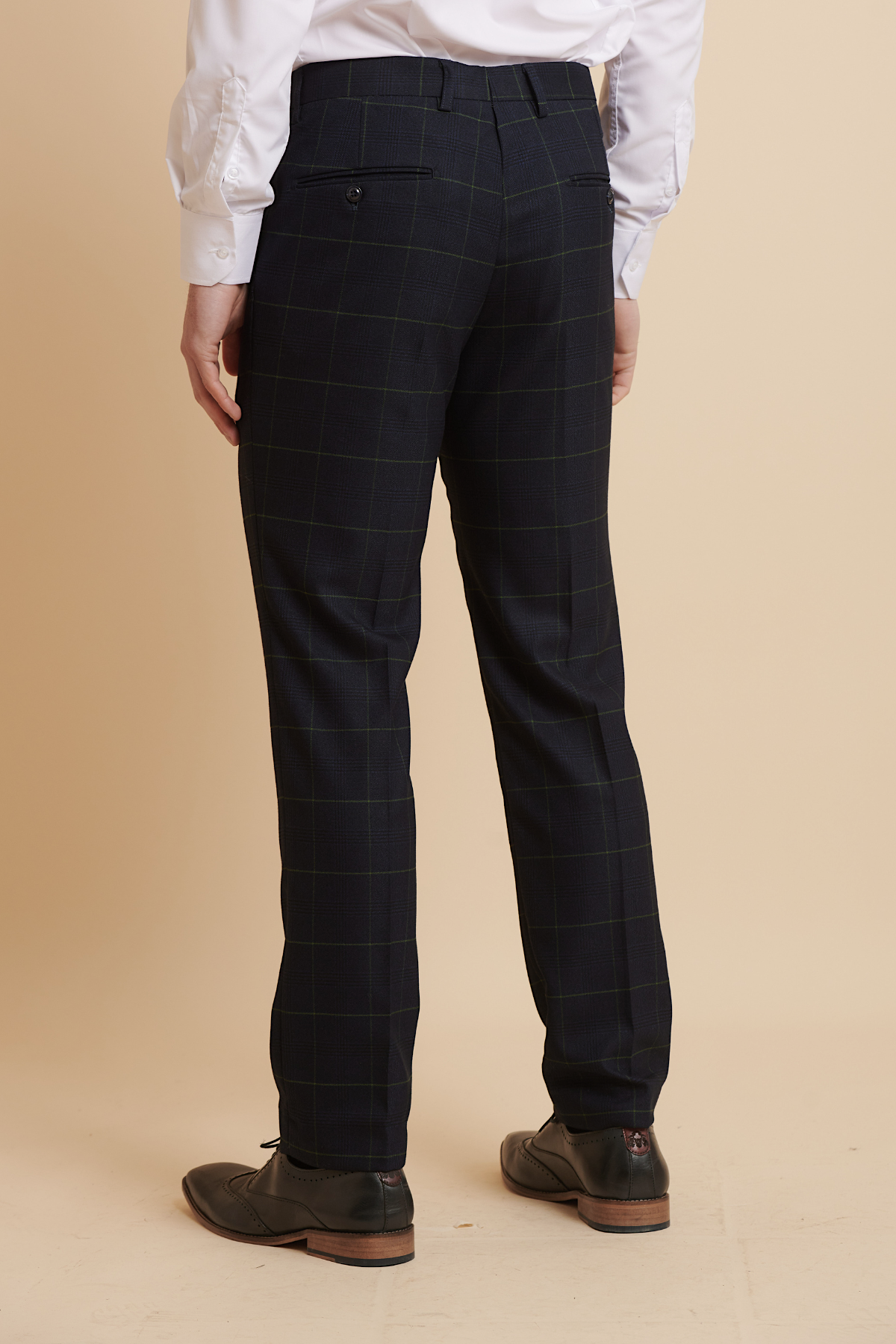 A person in Marc Darcy’s EDINSON Navy Green Check Trousers, a white dress shirt, and brown dress shoes stands with their back to the camera, highlighting the trousers against a beige background.