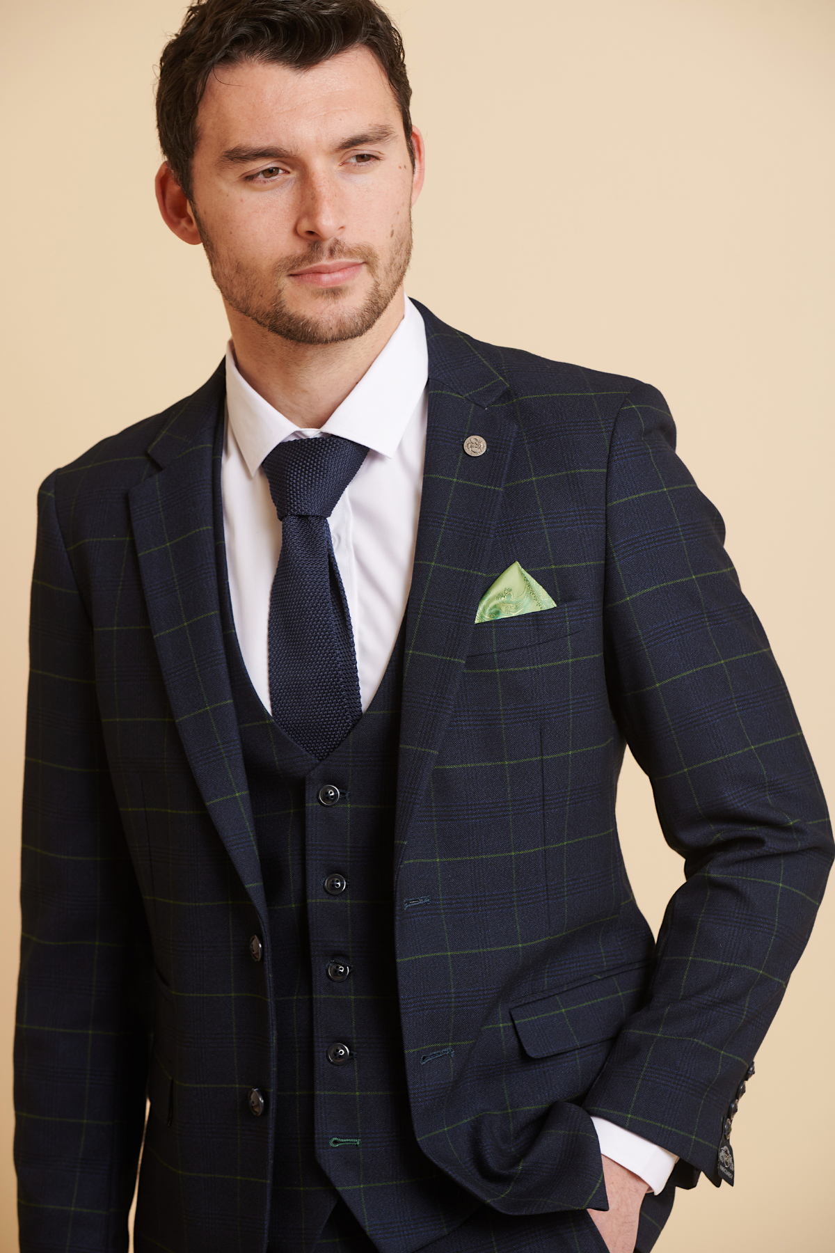 James Tarkowski wears the Marc Darcy Edinson Navy Green Check Suit from The Everton Collection, styled with a white shirt, navy tie, and green pocket square, standing with one hand in his pocket against a beige background.