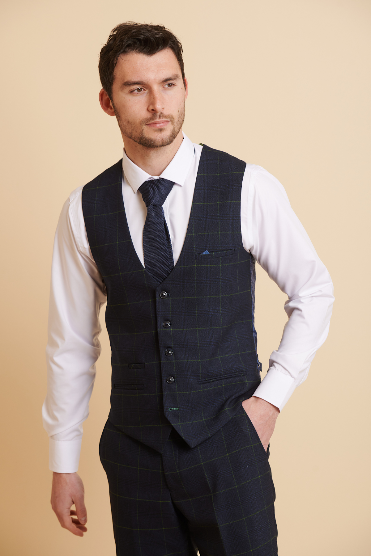 A man in a Marc Darcy EDINSON navy green check single breasted waistcoat with matching trousers, white dress shirt, and dark tie stands against a beige background, one hand in his pocket, looking to the side.