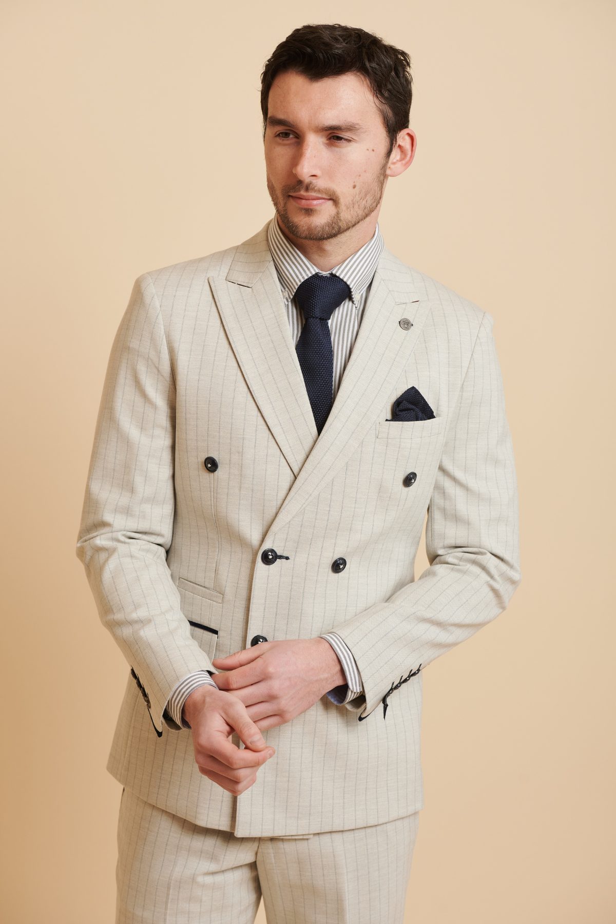 A man in the Marc Darcy Everton Collection Beto in GRANT Stone pinstripe double-breasted suit stands against a beige background, hands loosely clasped, looking slightly to the side.