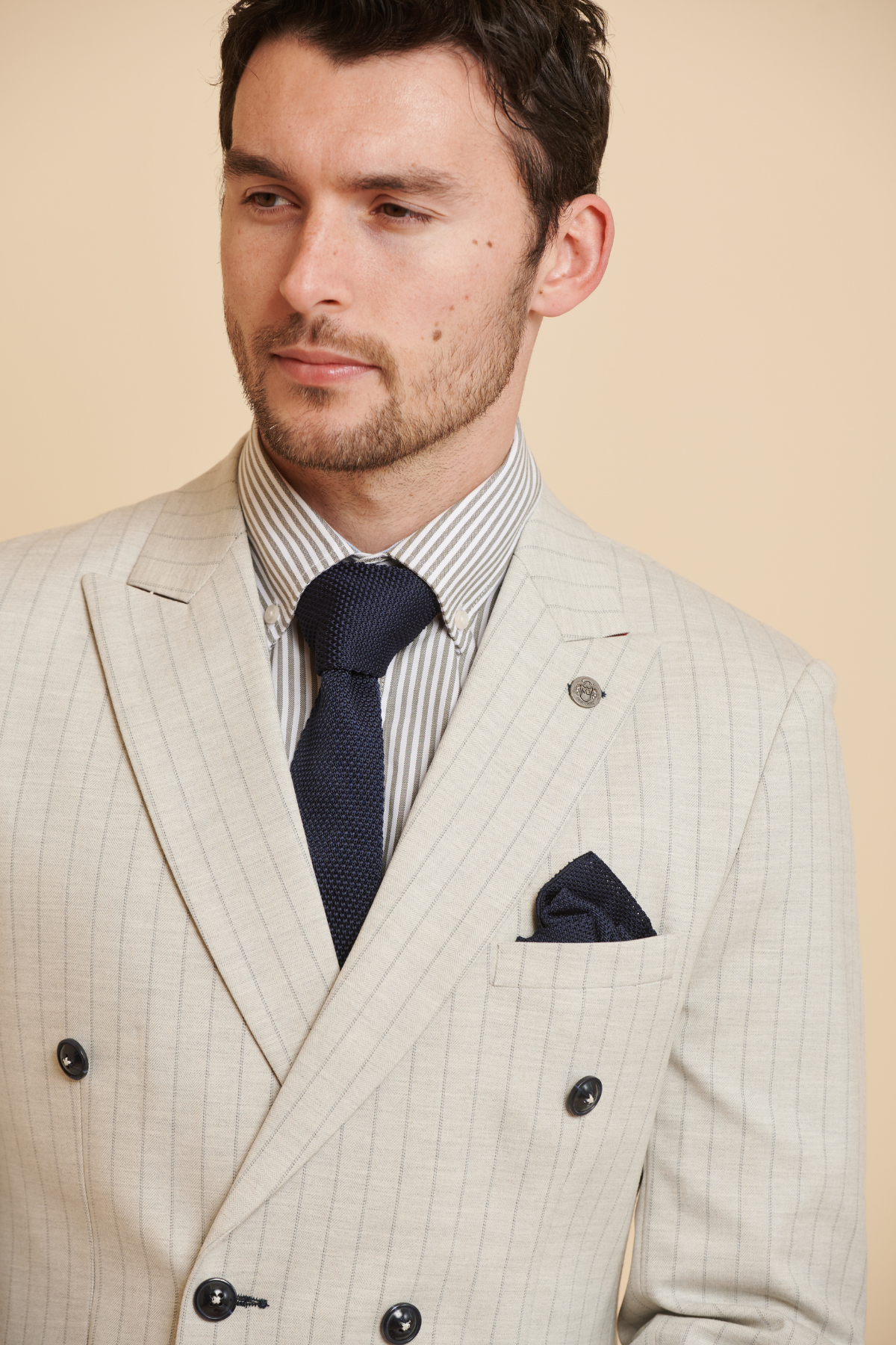 A man in the Marc Darcy Everton Collection | Beto in GRANT Stone Pinstripe Double Breasted Suit with a matching pocket square, navy tie, and gray striped shirt. He has short brown hair and a trimmed beard, looking slightly to the side.