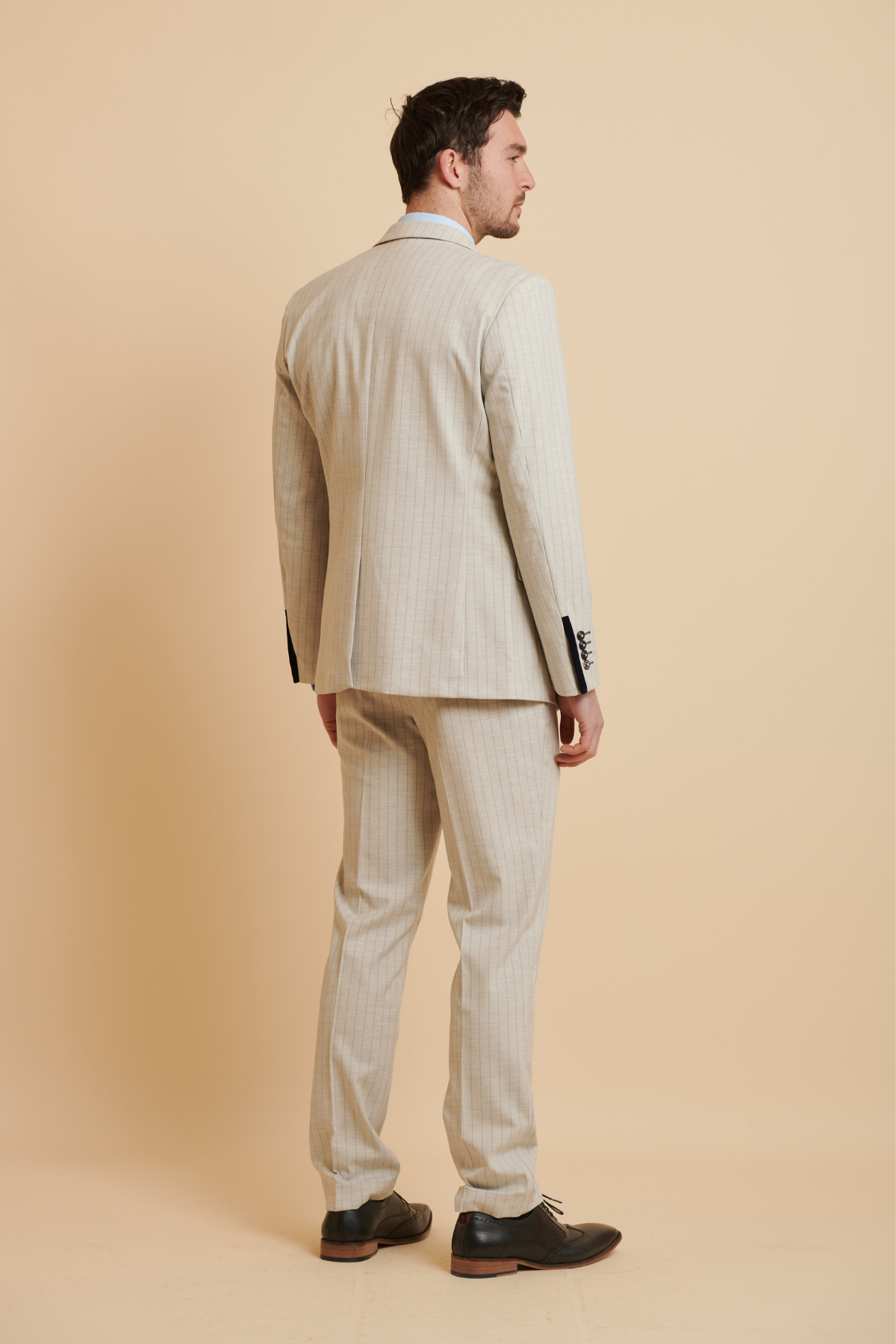 A man wears the Marc Darcy Everton Collection Beto in Grant Stone Pinstripe Double Breasted Slim Fit Three Piece Suit with black shoes, standing facing away from the camera against a plain beige background.