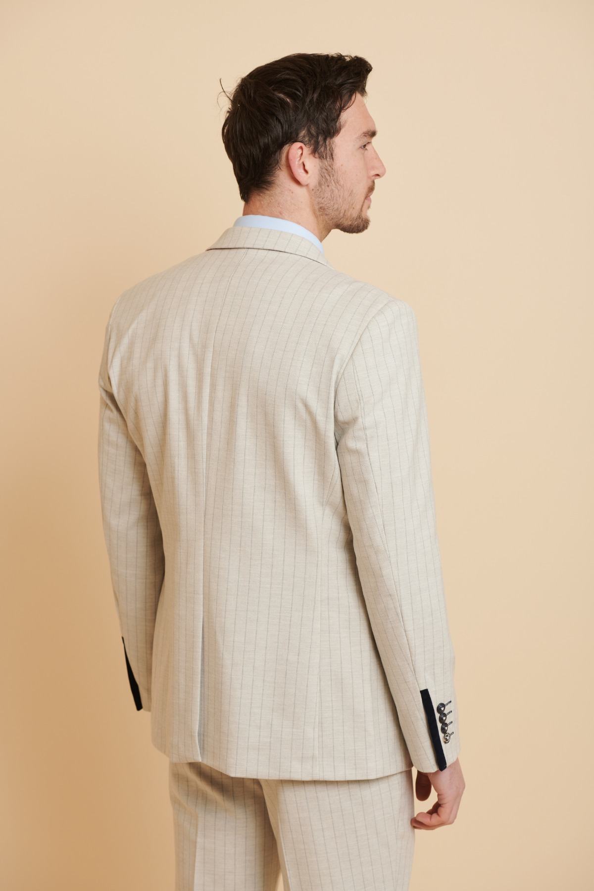 A man is seen from behind in Marc Darcy's The Everton Collection | Beto In GRANT Stone Pinstripe Double Breasted Suit, paired with a light blue shirt; this slim fit suit features dark sleeve buttons for a modern, tailored look.