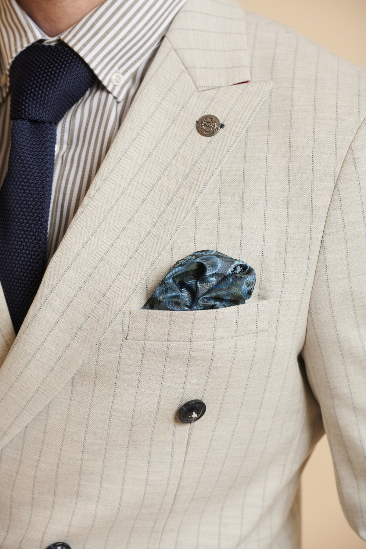 A person in Marc Darcy's The Everton Collection | Beto In GRANT Stone Pinstripe Double Breasted Suit, with striped shirt, navy tie, blue patterned pocket square, and lapel decorative buttons.