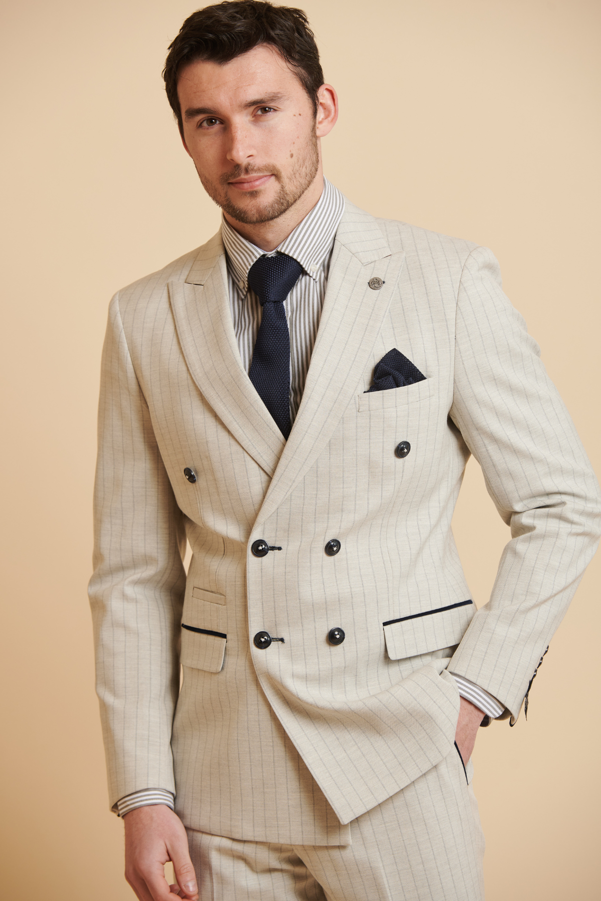 A man in The Everton Collection | Beto In GRANT Stone Pinstripe Double Breasted Suit by Marc Darcy, paired with a striped shirt, dark tie, and pocket square, stands confidently against a beige background.
