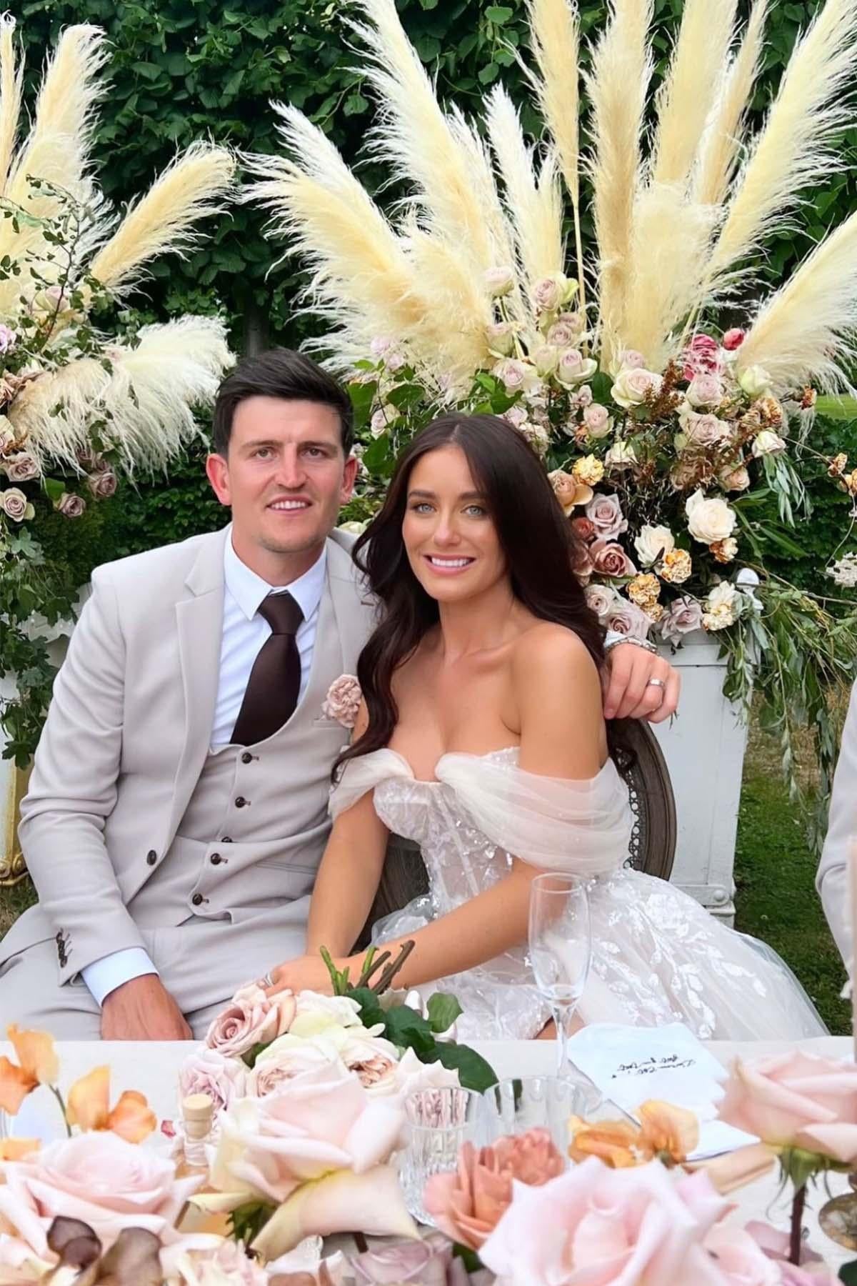 A couple sits at a decorated outdoor table with blush roses and pampas grass. The man wears the Marc Darcy Harry Maguire Wedding Suit HM5 Stone Three Piece Suit; the woman in an off-shoulder white dress smiles at the camera.