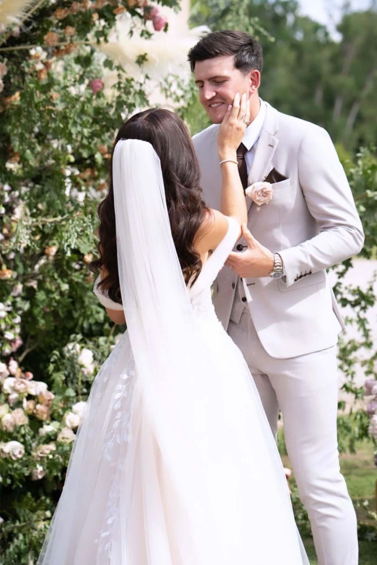A bride in a white dress and veil gently touches her smiling groom, who is wearing the Marc Darcy Harry Maguire Wedding Suit - HM5 Stone Three Piece Suit, as they stand outdoors near lush greenery and flowers.