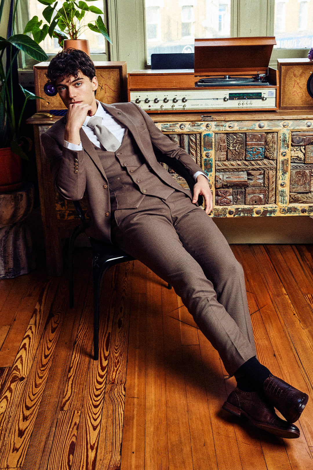A man wearing the Marc Darcy HM5 Tan Tailored Three Piece Suit sits casually in a stylish room with wooden floors, a vintage radio, and large windows, resting his chin on his hand and looking thoughtfully at the camera.