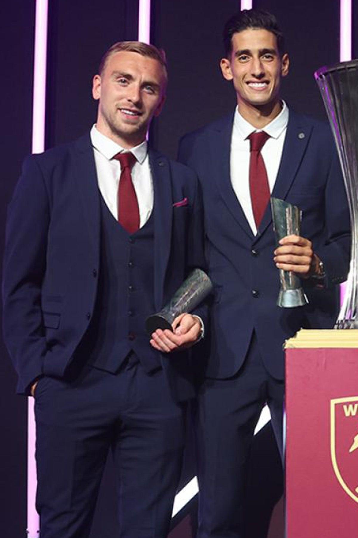 West Ham United footballer Jarrod Bowen, in a Marc Darcy Bromley Navy Check Suit, stands beside another man in matching attire. Both hold glass trophies before a large silver cup on a podium, with vertical neon lights illuminating the background.