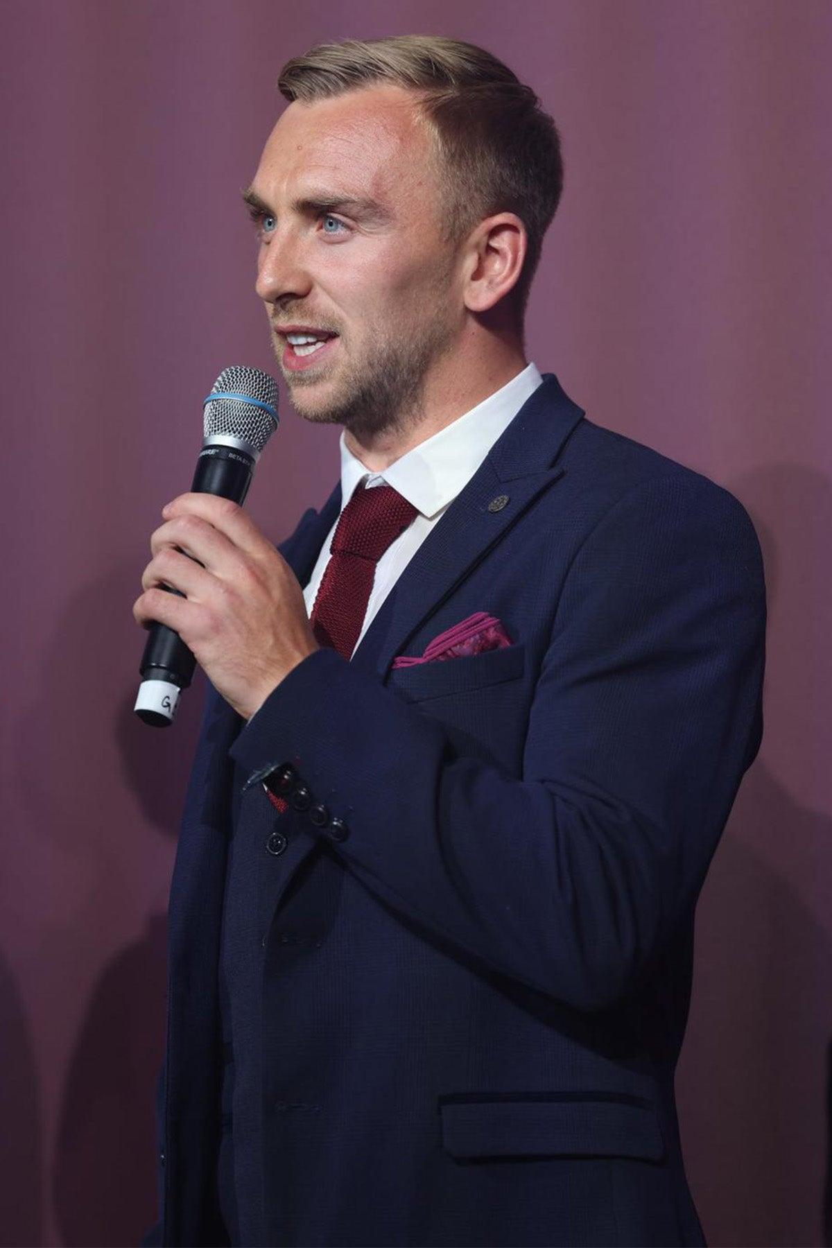 West Ham United footballer Jarrod Bowen wears a Bromley Navy Check Suit by Marc Darcy, paired with a burgundy tie and pocket square, as he holds a microphone and speaks against a plain maroon background.