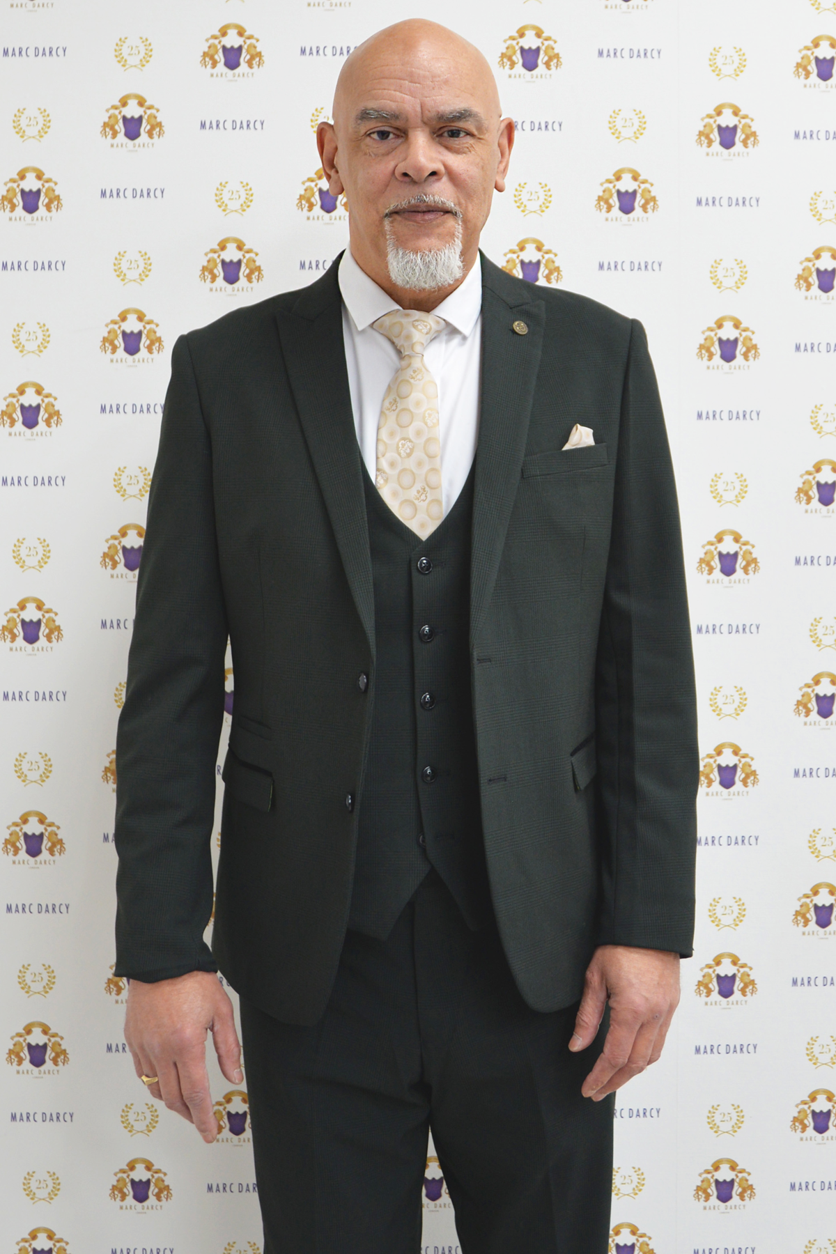Former Wolverhampton Wanderers footballer George Berry sports a Marc Darcy Bromley olive green 3-piece suit, white shirt, and patterned tie in front of a branded backdrop.