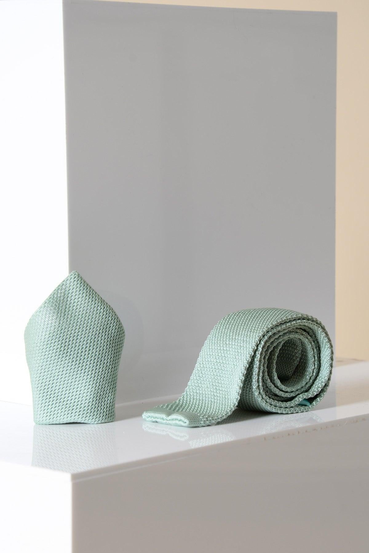 The Accessory KT - Green Knitted Tie & Pocket Square are shown on a glossy white surface with a minimalistic backdrop, emphasizing the elegant texture and design of the sage green set.