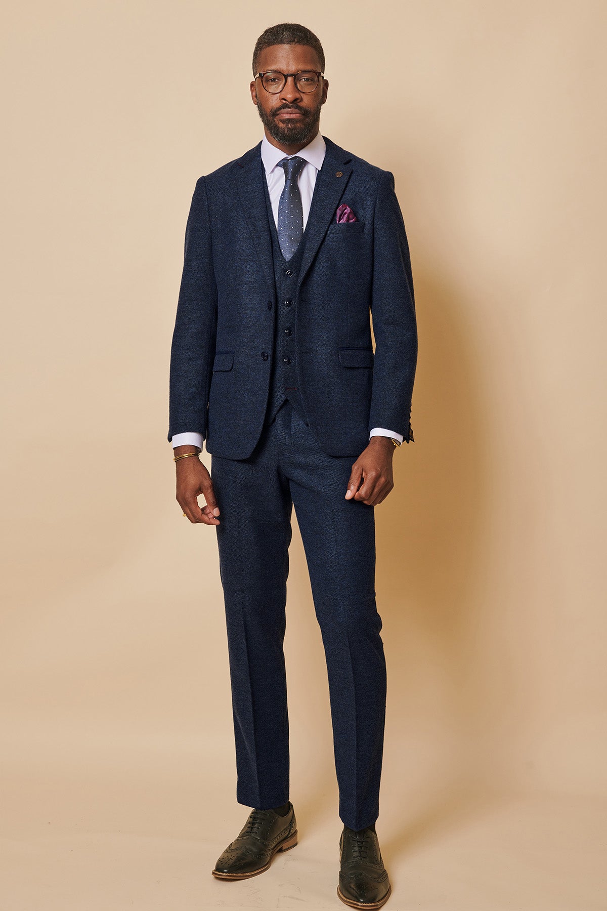 A man wearing glasses poses against a beige background in the marcdarcy MARLOW navy tweed three-piece suit, styled slim fit, with a white shirt, blue patterned tie, purple pocket square, and brown shoes.
