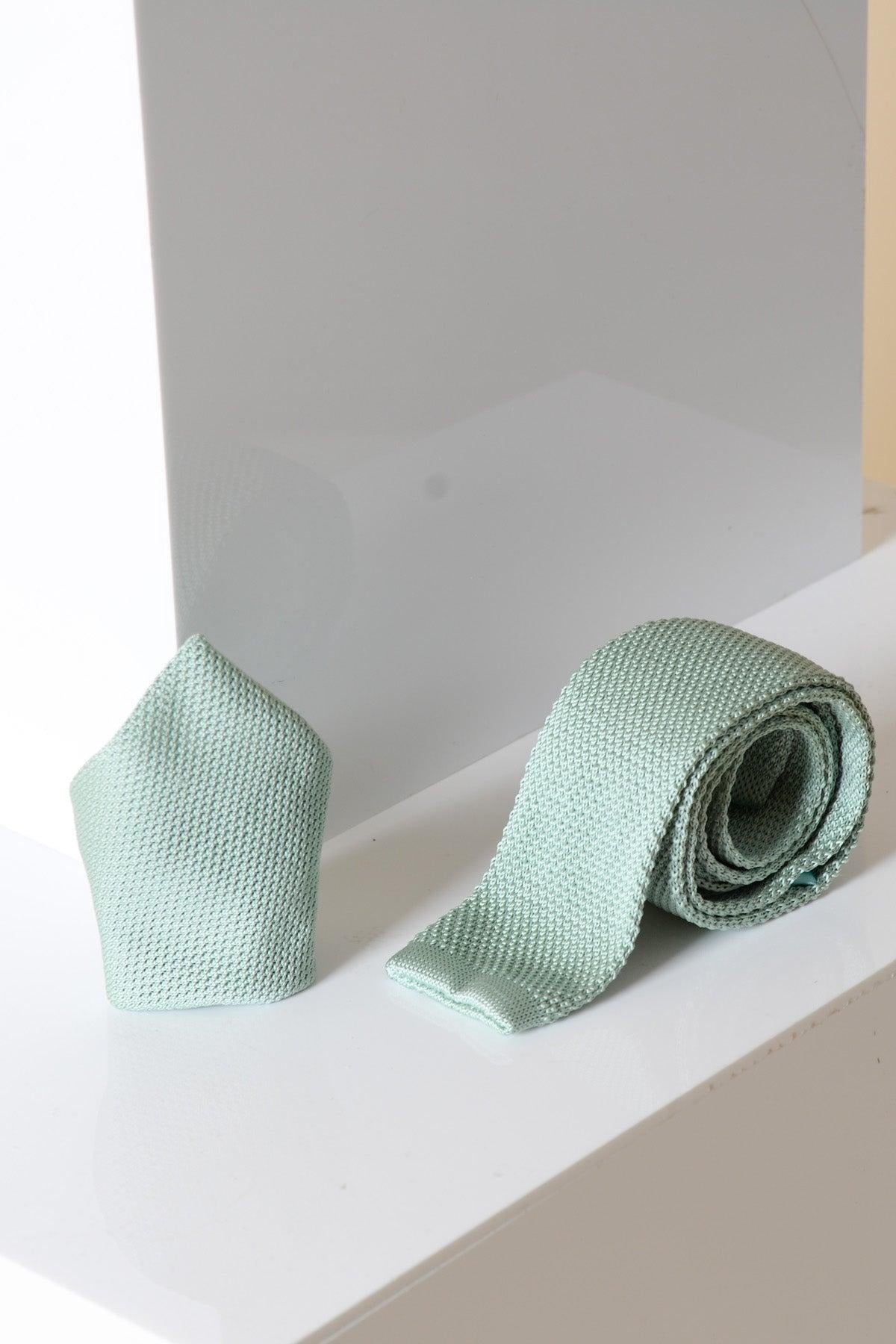 The Accessory KT - Green Knitted Tie & Pocket Square, featuring a textured sage green design, is displayed rolled up with the matching pocket square on a glossy white surface against a soft white and gray background.