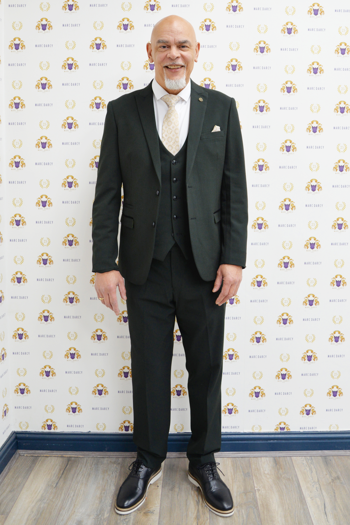 Former Wolverhampton Wanderers footballer George Berry smiles in a Marc Darcy Bromley Olive Green Suit, featuring a vest and cream tie, standing before a wall with a repeating “Blue Daisy” logo pattern on a wood floor.