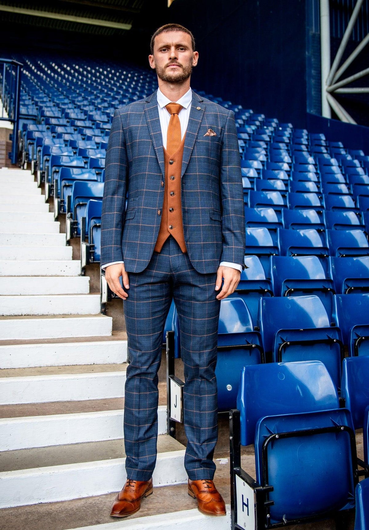 A man in marcdarcy’s WBA Collection Jenson Marine Suit with Kelvin Tan Waistcoat, as worn by John Swift, stands before empty blue stadium seats and white stairs, looking directly at the camera.