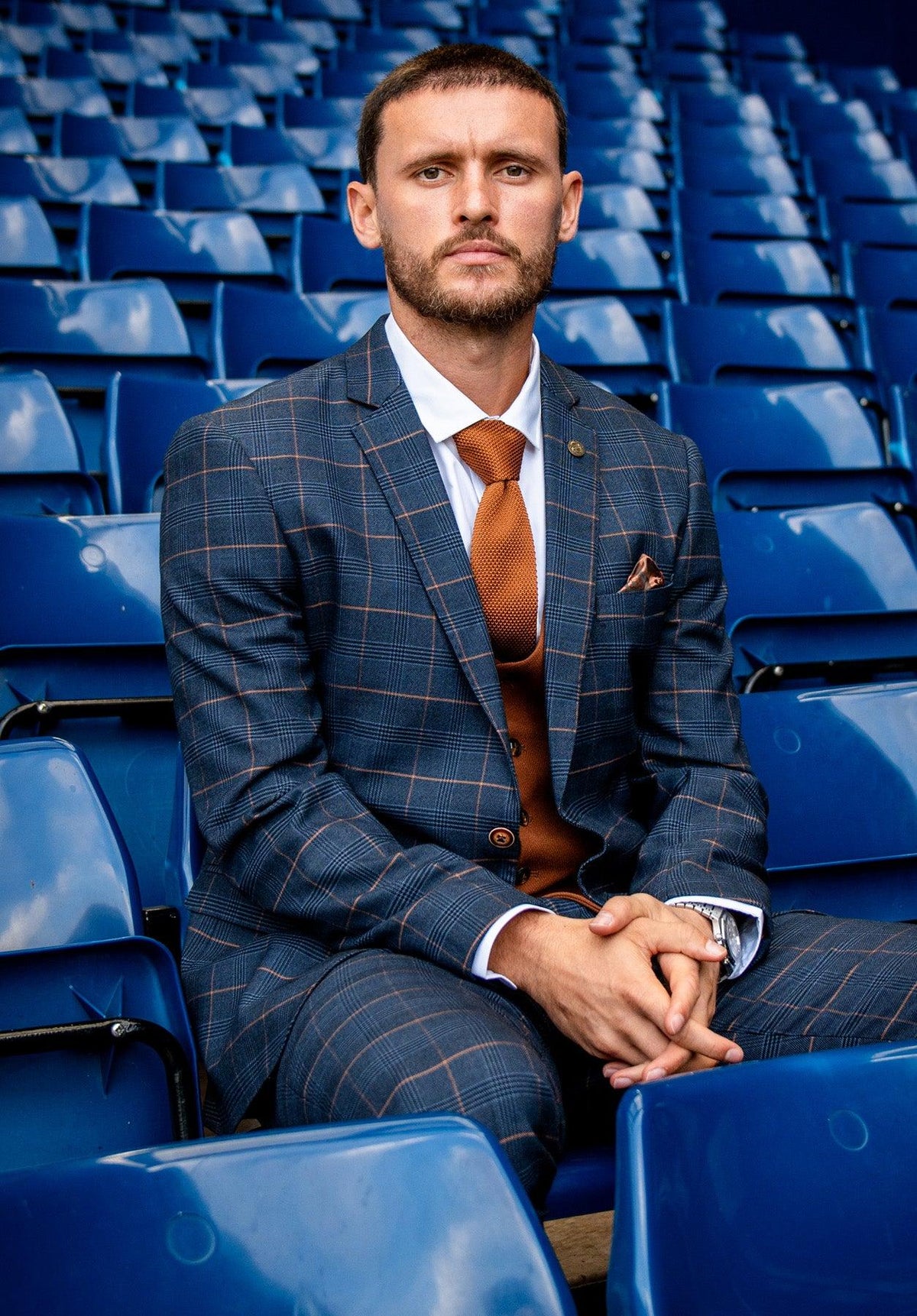 A man wears the marcdarcy WBA Collection – Jenson Marine Suit with Kelvin Tan Waistcoat, as worn by John Swift, paired with an orange tie, sitting seriously on blue stadium seats with hands clasped.